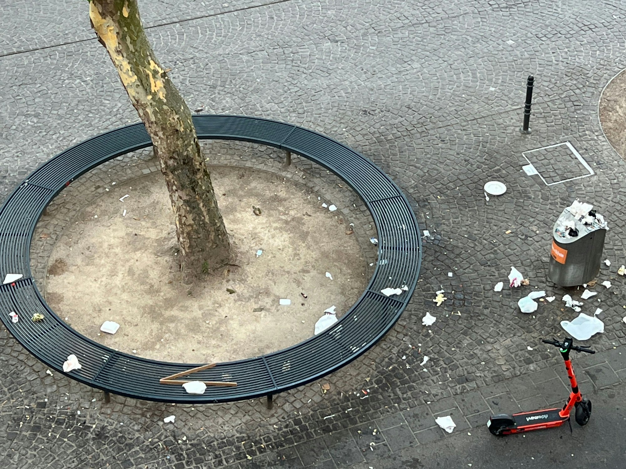 Auf einer runden Sitzbank liegt Müll, ebenso auf dem Boden rund um einen Mülleimer.