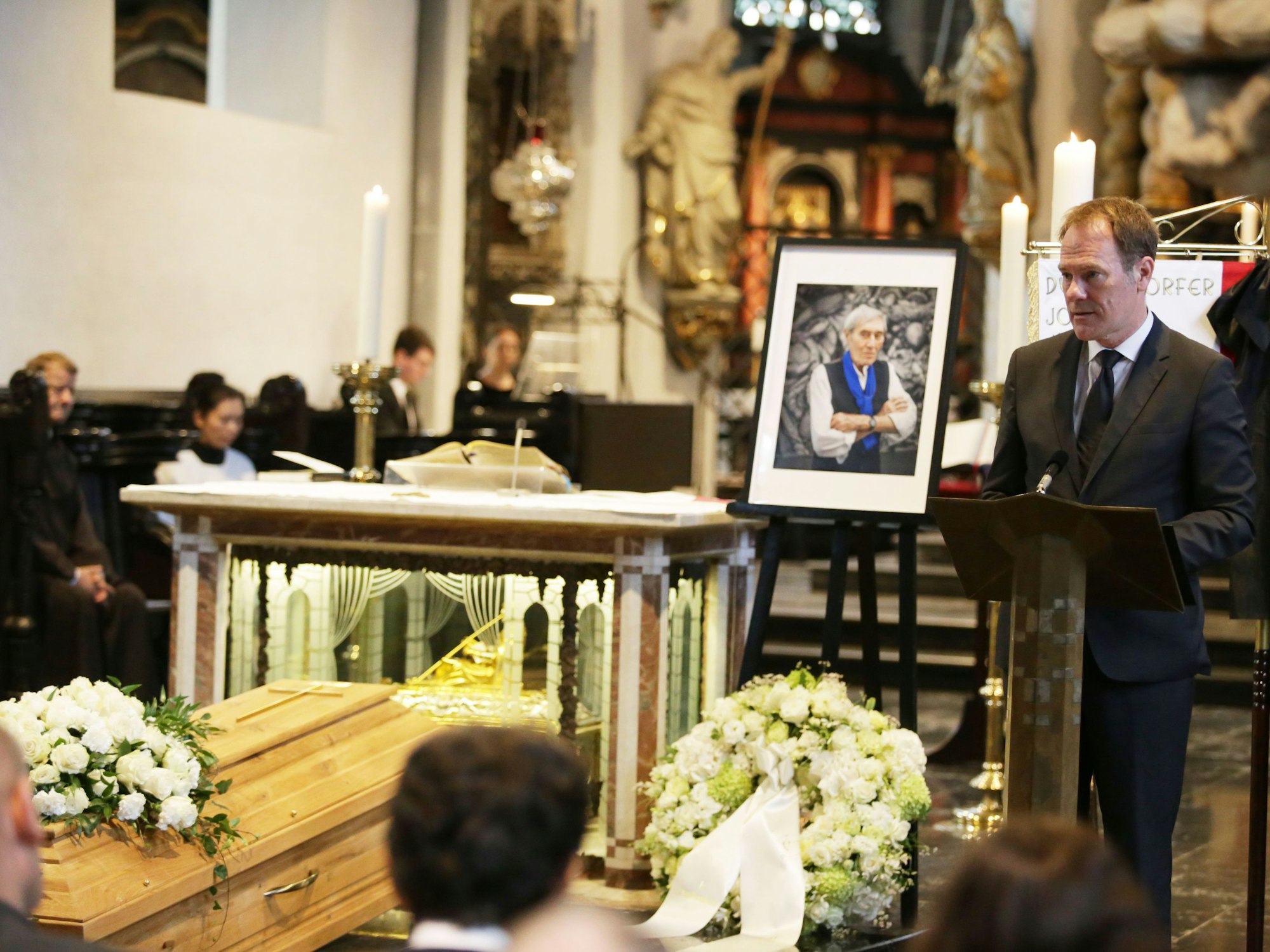 Ein Mann steht in einer Kirche an einem Rednerpult, neben ihm ein Sarg mit Kränzen.