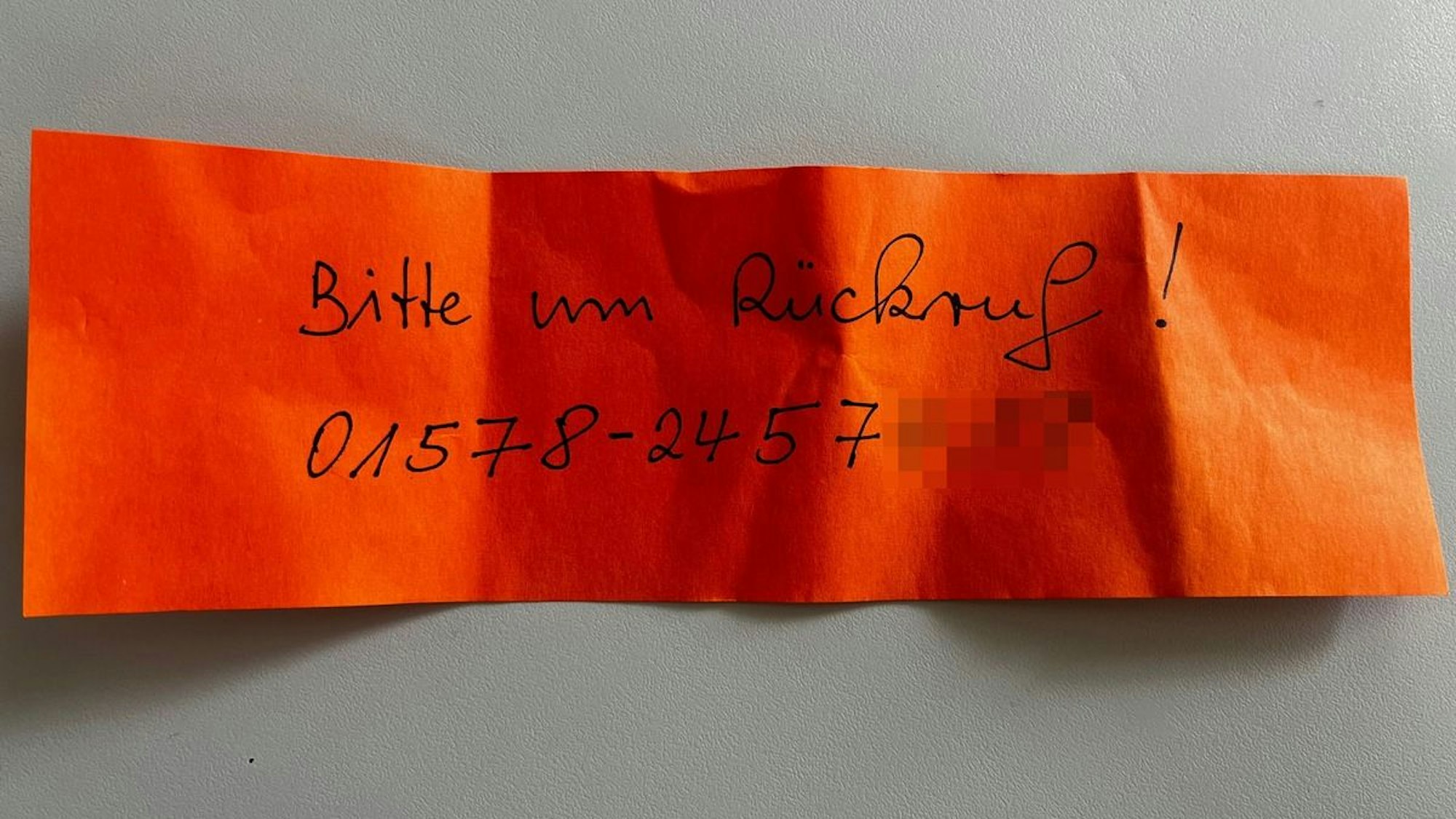 Ein oranger Zettel mit einer Telefonnummer klemmt an einem Auto.