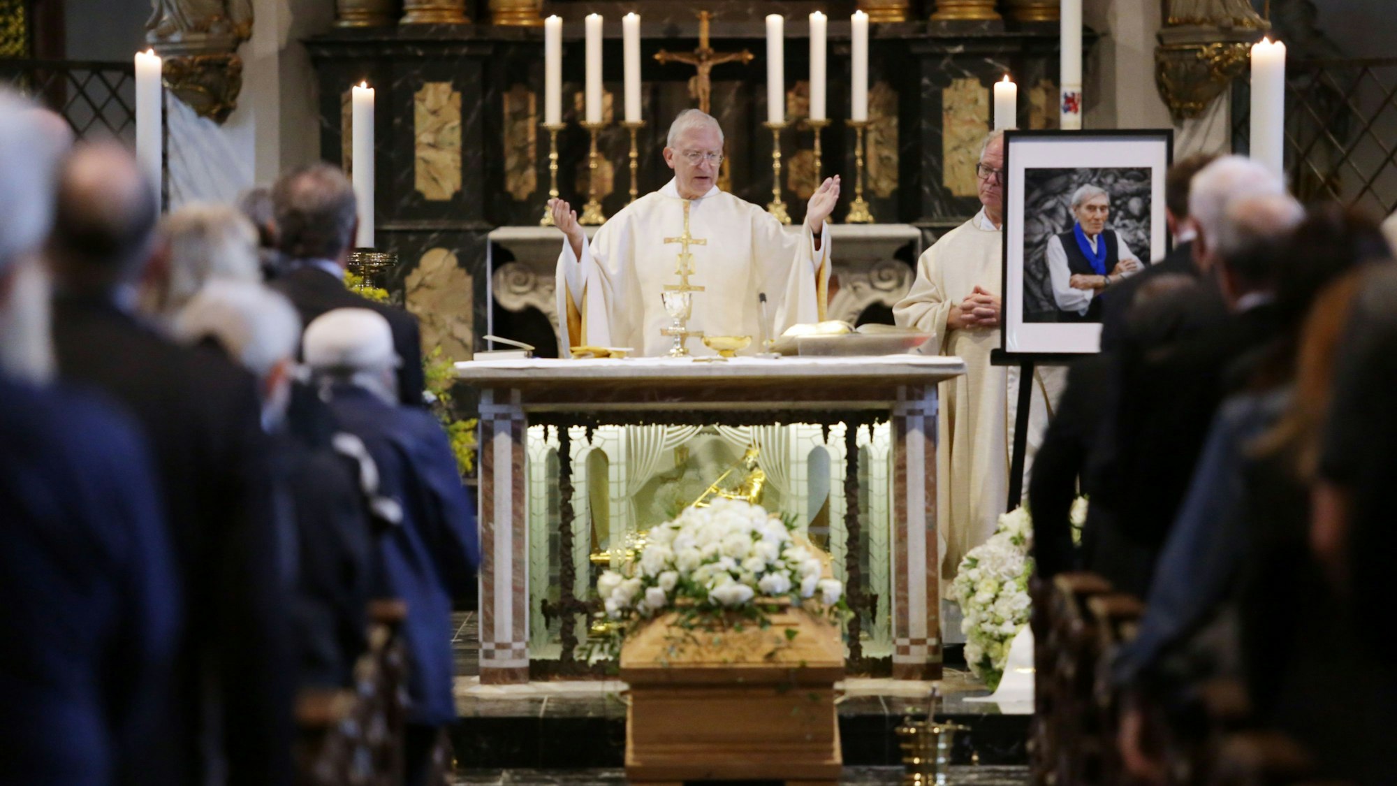 Ein Pfarrer steht in einer mit Menschen gefüllten Kirche hinter dem Altar, davor steht ein Sarg mit Blumen.