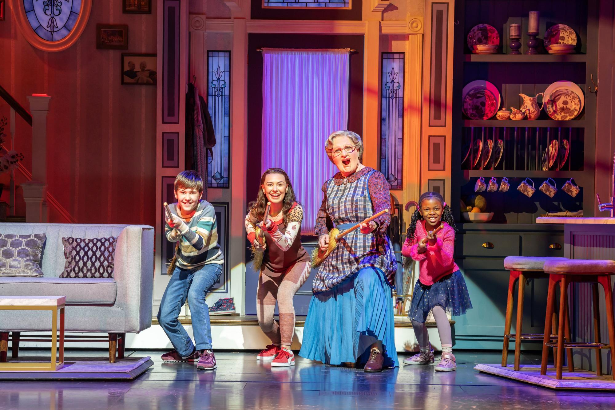 Musicalszene mit Mrs. Doubtfire und drei Kindern, die lachend mit Besen Gitarre spielen.
