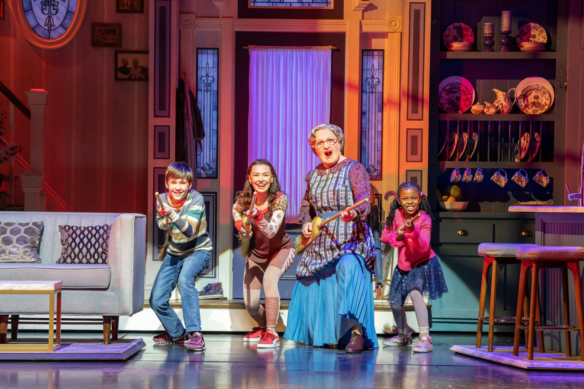 Musicalszene mit Mrs. Doubtfire und drei Kindern, die lachend mit Besen Gitarre spielen.
