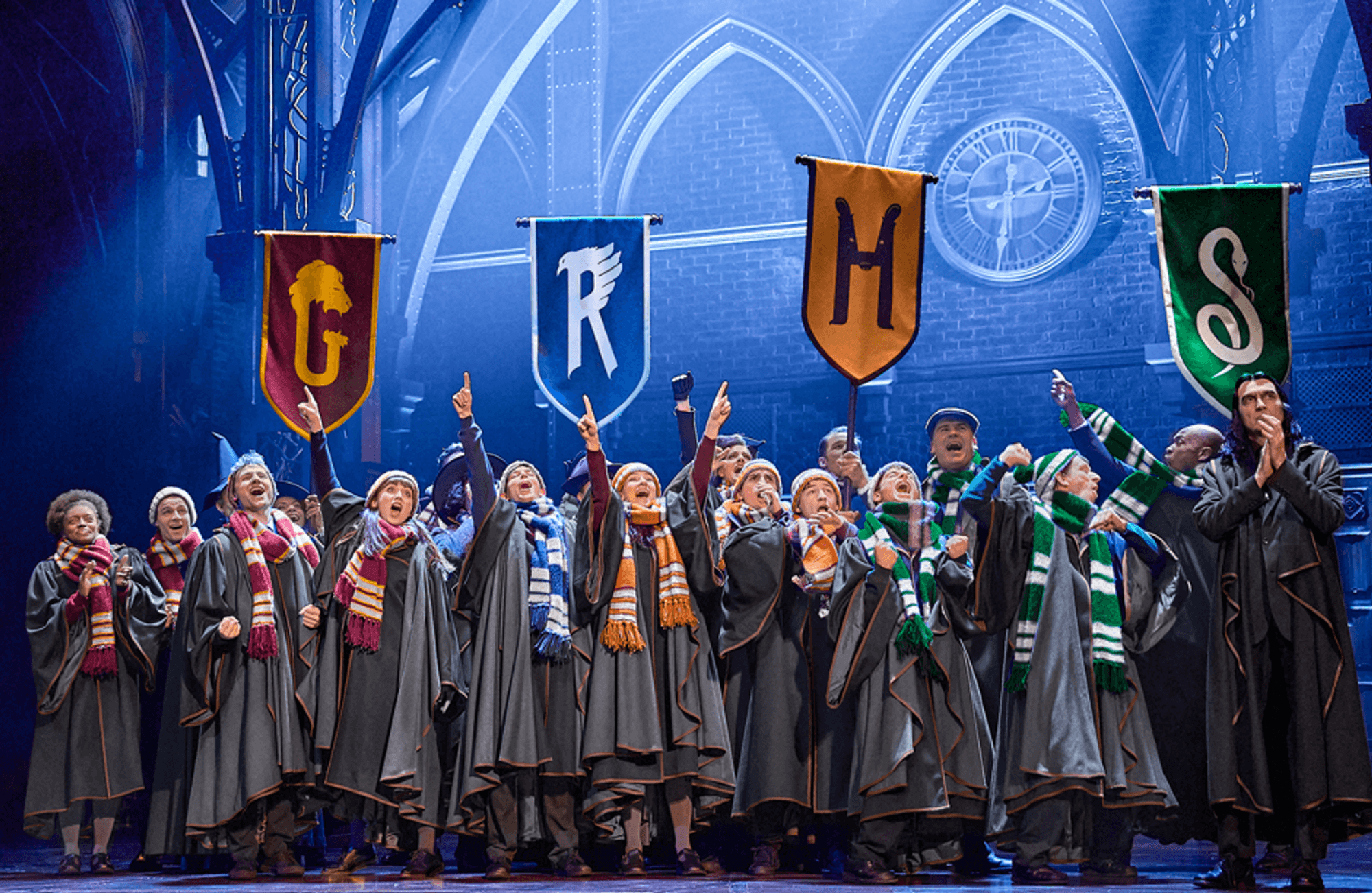 In Hogwarts-Roben und mit bunten Schals stehen Schüler jubelnd unter Fahnen der vier Häuser Gryffindor, Ravenclaw, Hufflepuff und Slytherin auf der Bühne.