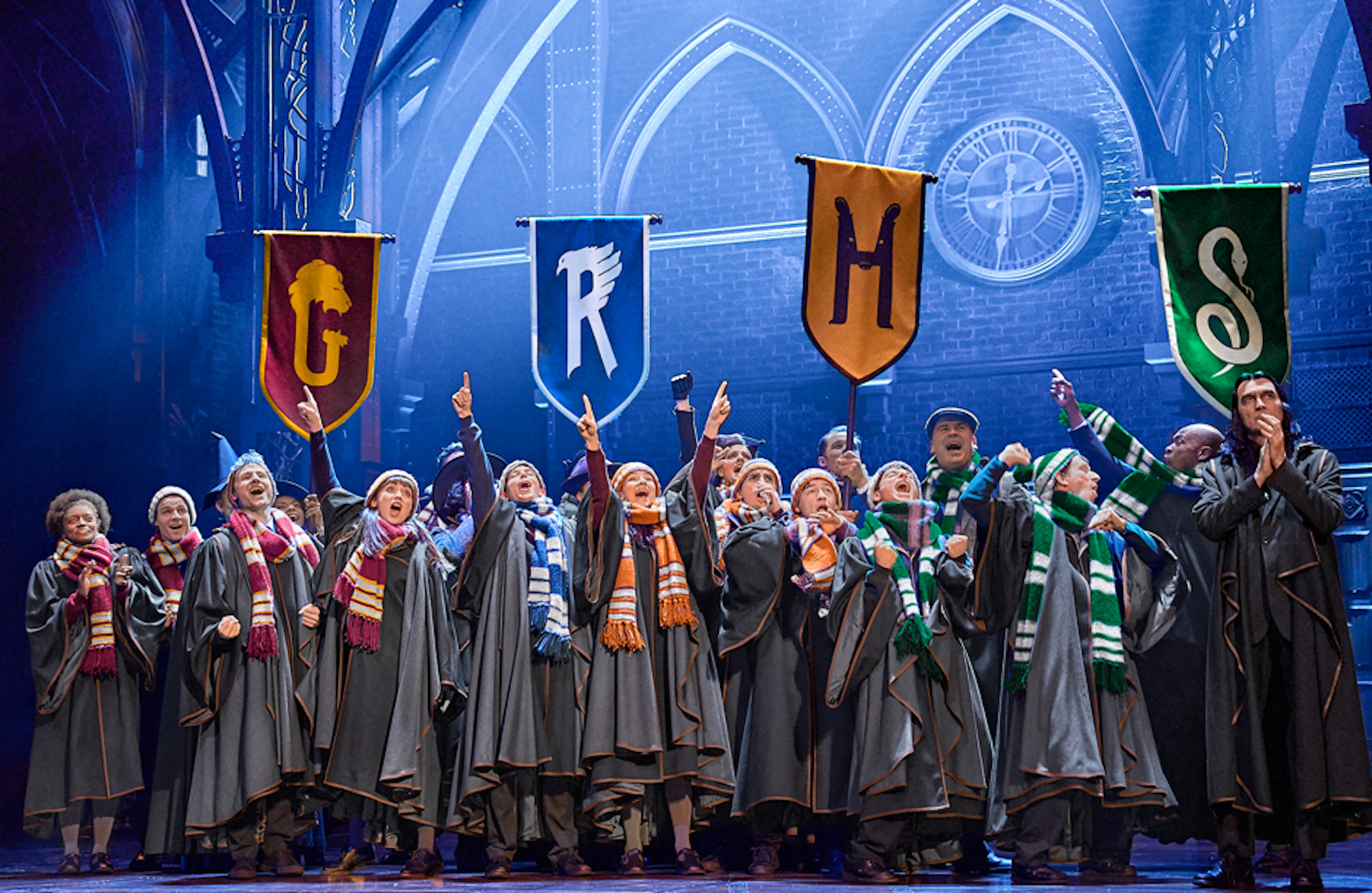 In Hogwarts-Roben und mit bunten Schals stehen Schüler jubelnd unter Fahnen der vier Häuser Gryffindor, Ravenclaw, Hufflepuff und Slytherin auf der Bühne.