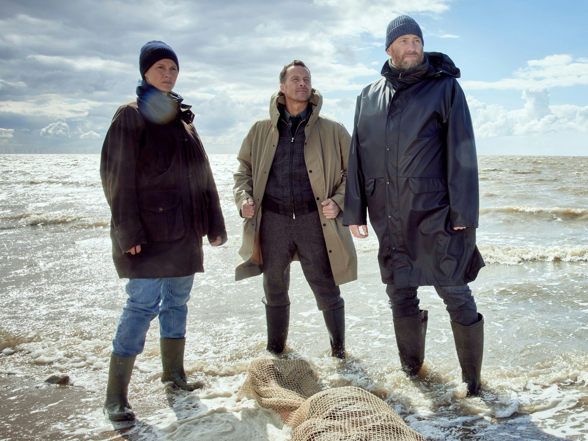 Drei Personen in Gummistiefeln stehen im Watt und blicken in die Kamera.