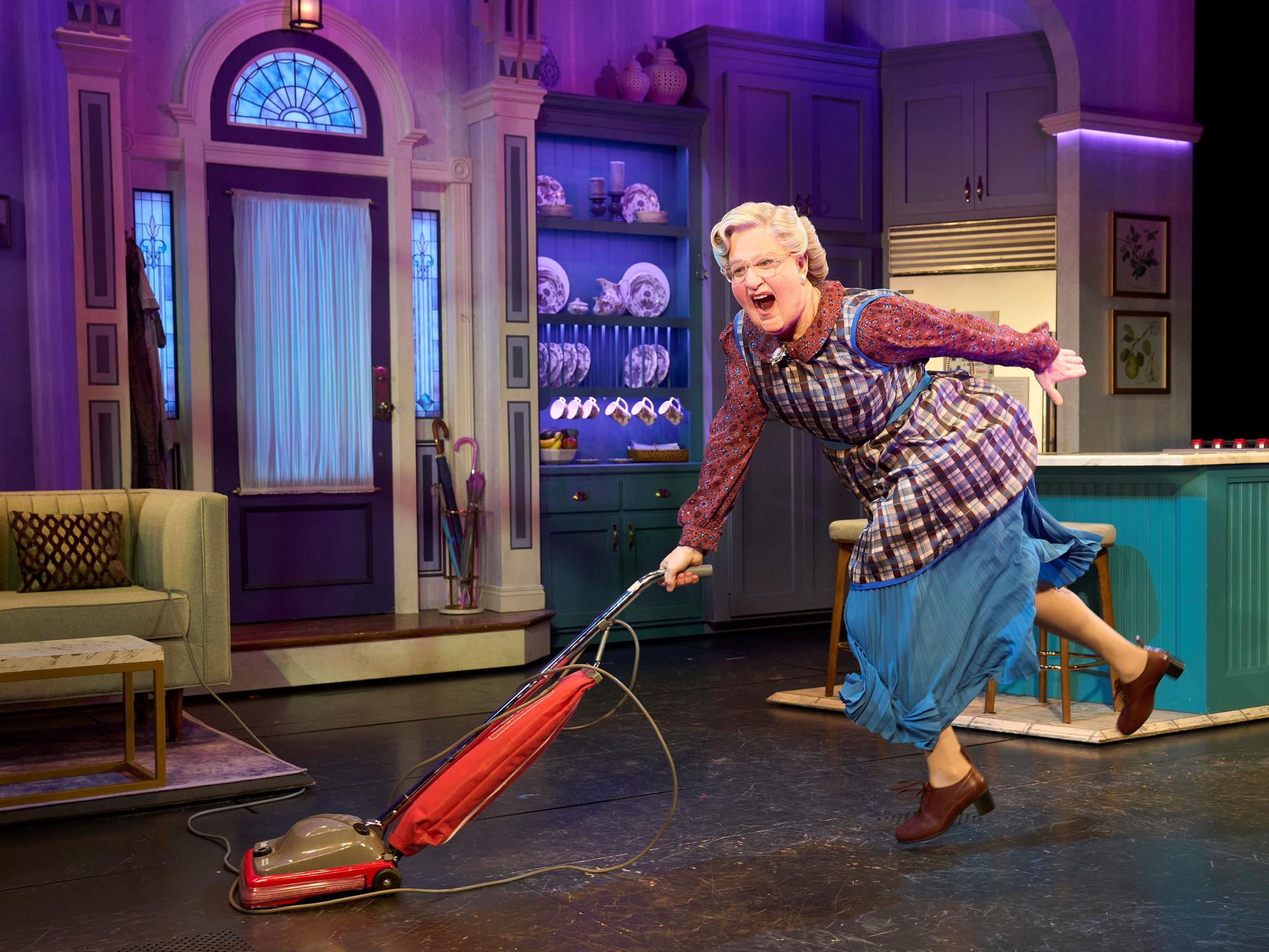 Mrs. Doubtfire tanzt mit einem roten Staubsauger durch eine bunte Bühnenküche, voller Energie.