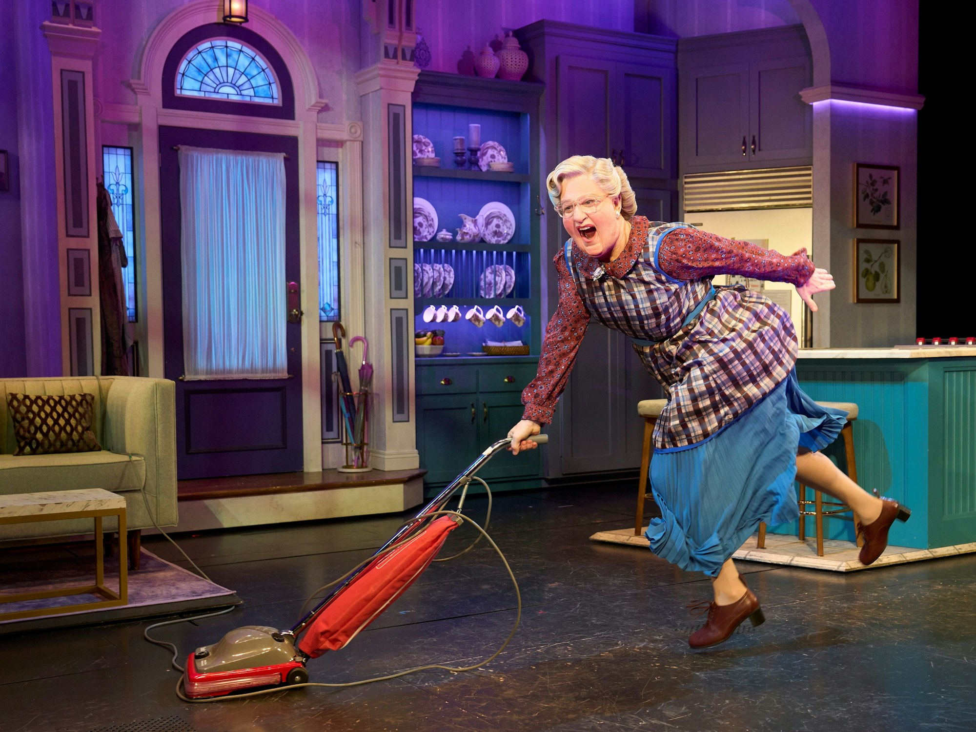 Mrs. Doubtfire tanzt mit einem roten Staubsauger durch eine bunte Bühnenküche, voller Energie.