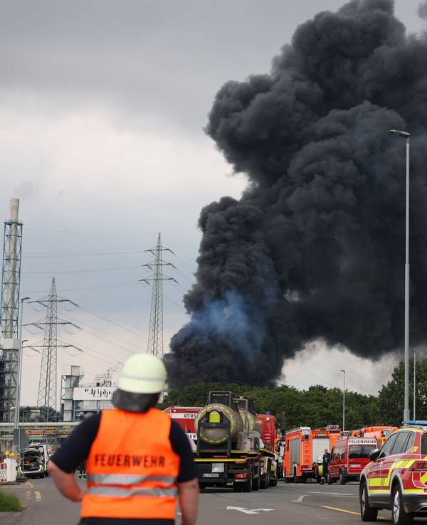 Einsatzfahrzeuge der Feuerwehr stehen unweit einer Zufahrt zum Chemiepark, über dem eine dunkle Rauchwolke aufsteigt.