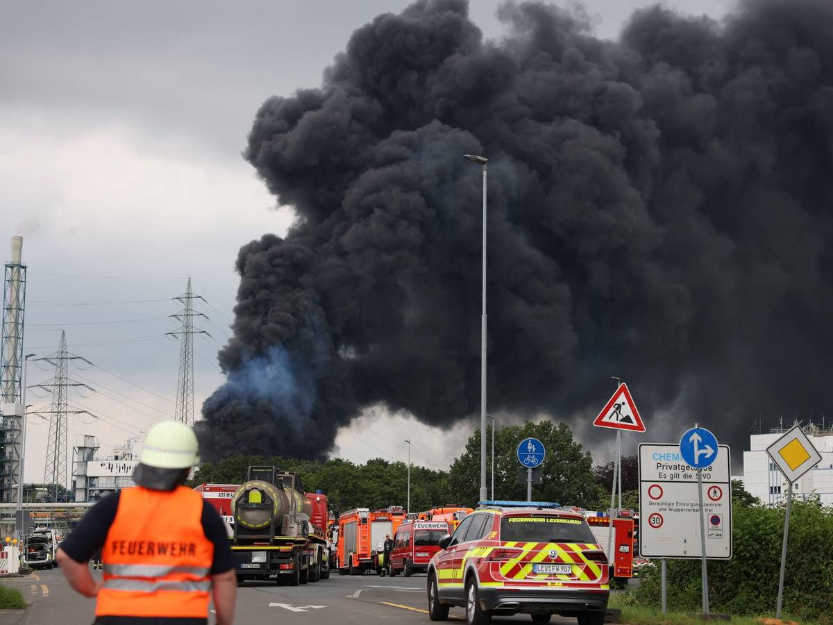 Einsatzfahrzeuge der Feuerwehr stehen unweit einer Zufahrt zum Chemiepark, über dem eine dunkle Rauchwolke aufsteigt.