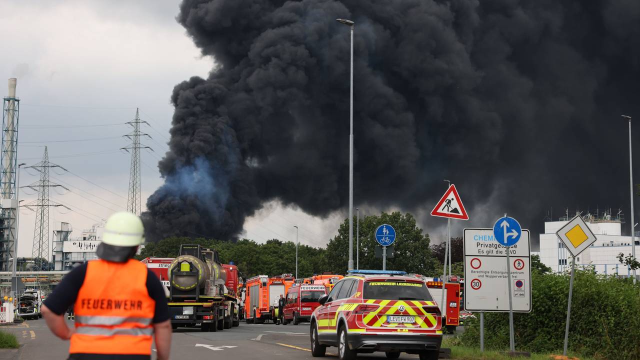 Einsatzfahrzeuge der Feuerwehr stehen unweit einer Zufahrt zum Chemiepark, über dem eine dunkle Rauchwolke aufsteigt.