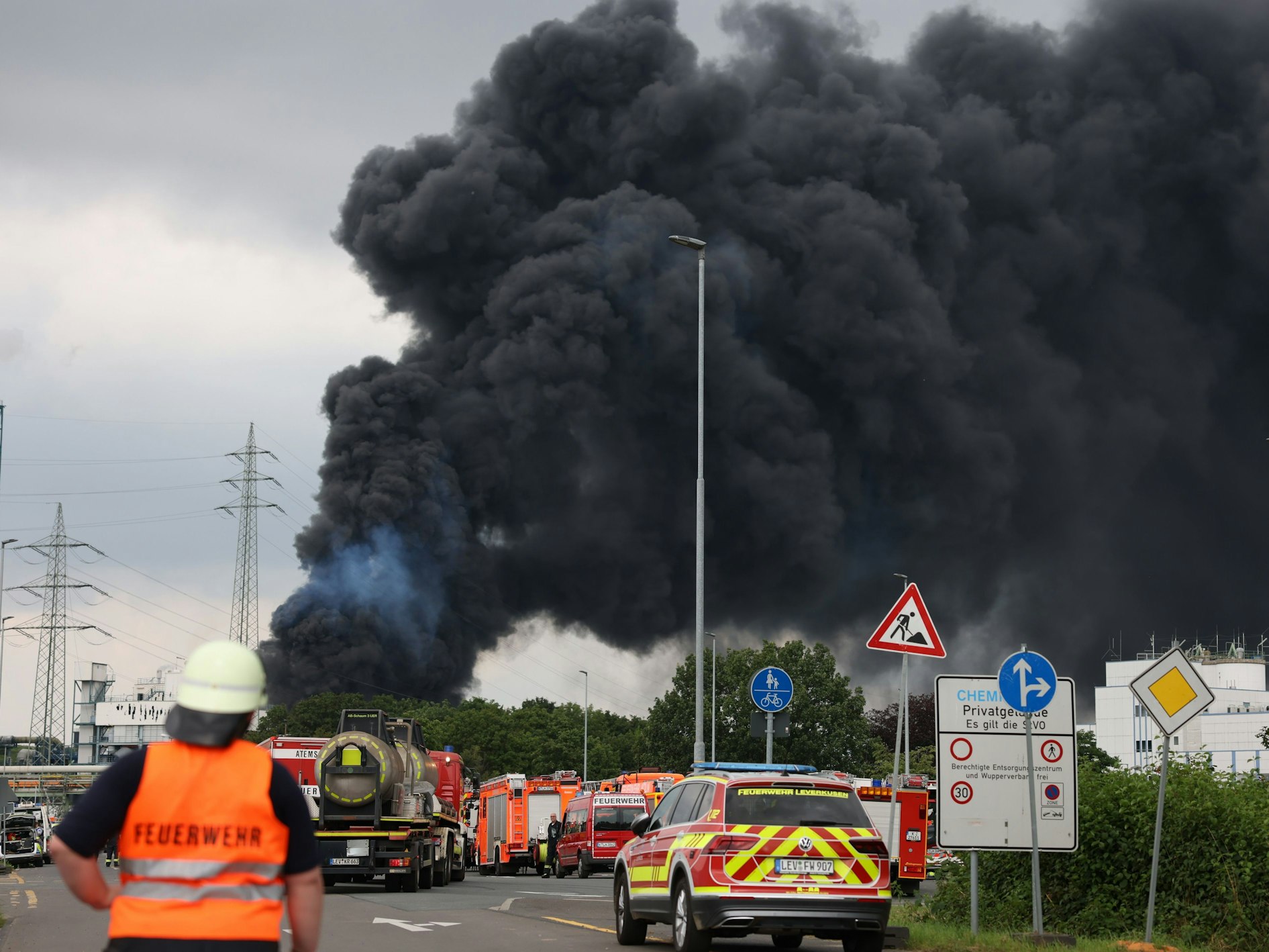 Einsatzfahrzeuge der Feuerwehr stehen unweit einer Zufahrt zum Chemiepark, über dem eine dunkle Rauchwolke aufsteigt.
