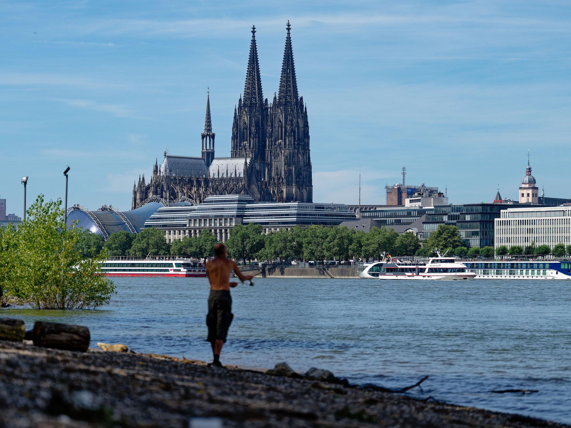 Ein Angler steht bei Hochsommerwetter am Rhein und angelt. Im Hintergrund der Kölner Dom