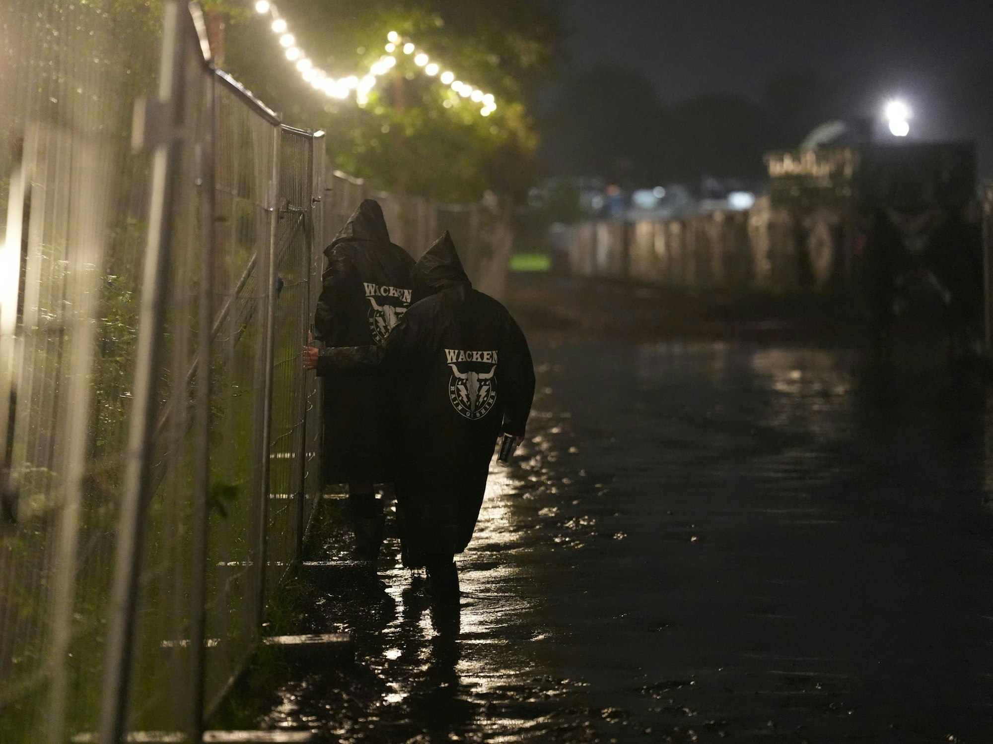 Metal-Fans gehen in der Nacht über einen überfluteten Weg auf dem Festivalgelände des Wacken Open Air (WOA).