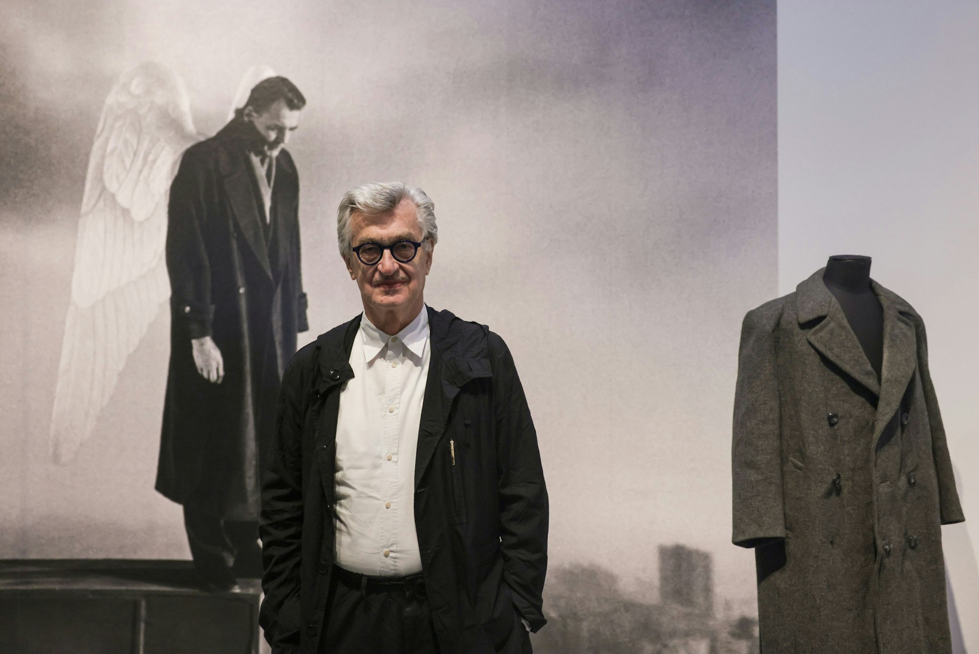 Wim Wenders steht vor einem Szenenfoto und dem Mantel aus dem Film „Der Himmel über Berlin".