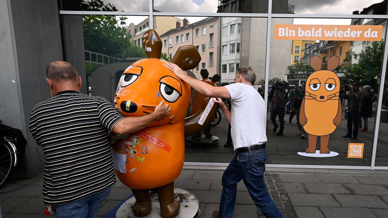 Zwei Männer schieben eine Maus-Figur auf einem Rollbrett weg.