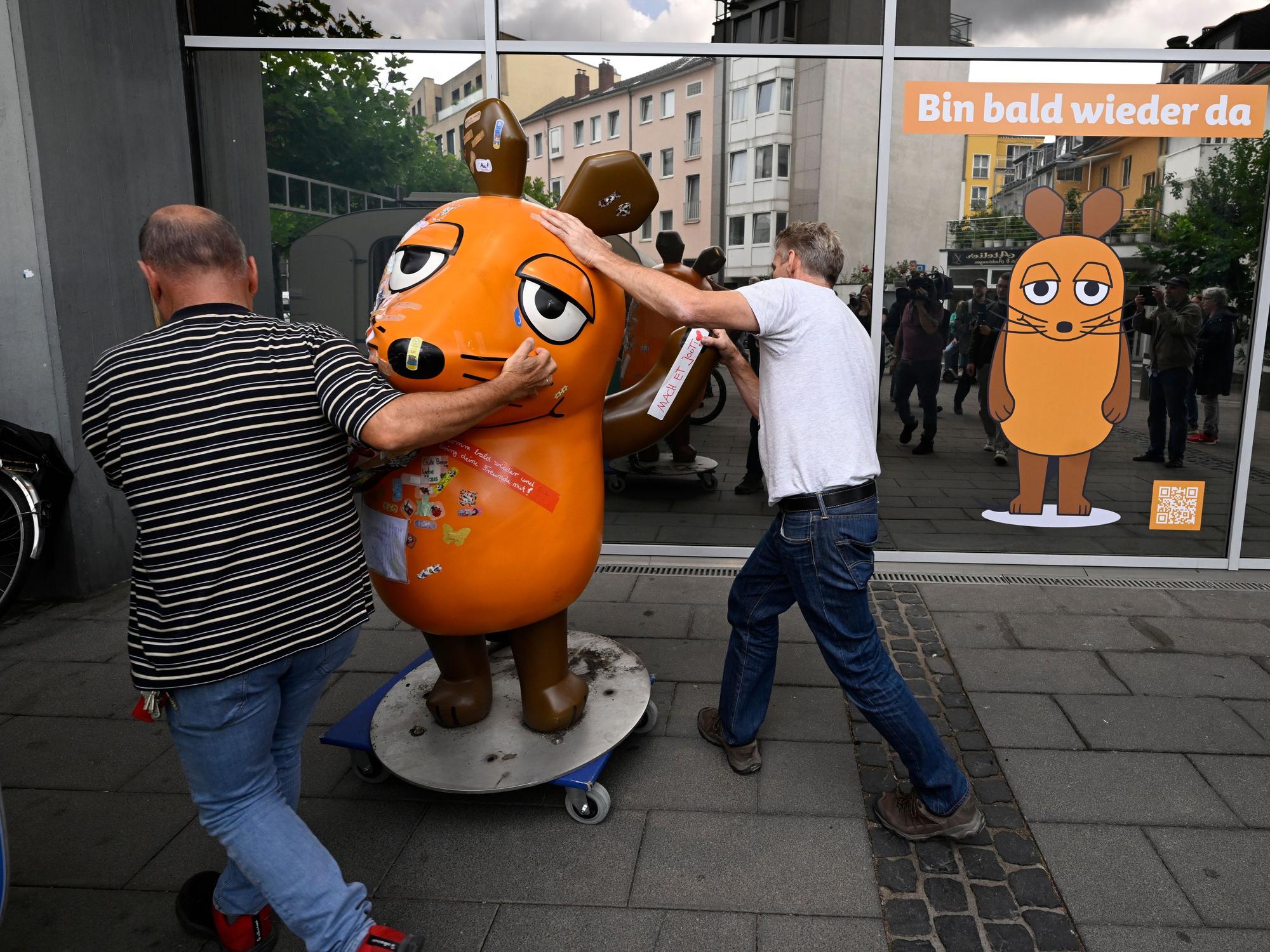 Zwei Männer schieben eine Maus-Figur auf einem Rollbrett weg.