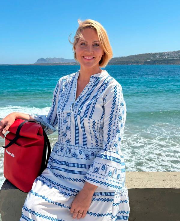 Eine blonde Frau in einem blau-weißen Kleid sitzt auf einer Mauer am Meer