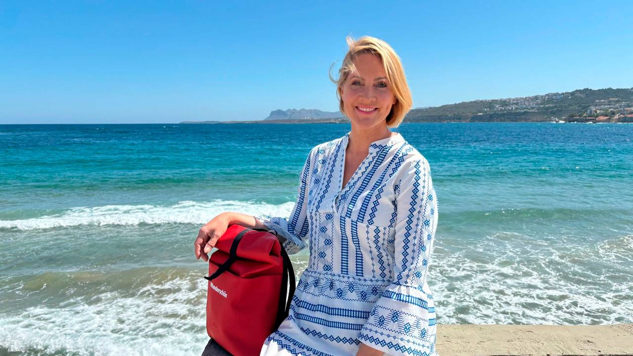 Eine blonde Frau in einem blau-weißen Kleid sitzt auf einer Mauer am Meer