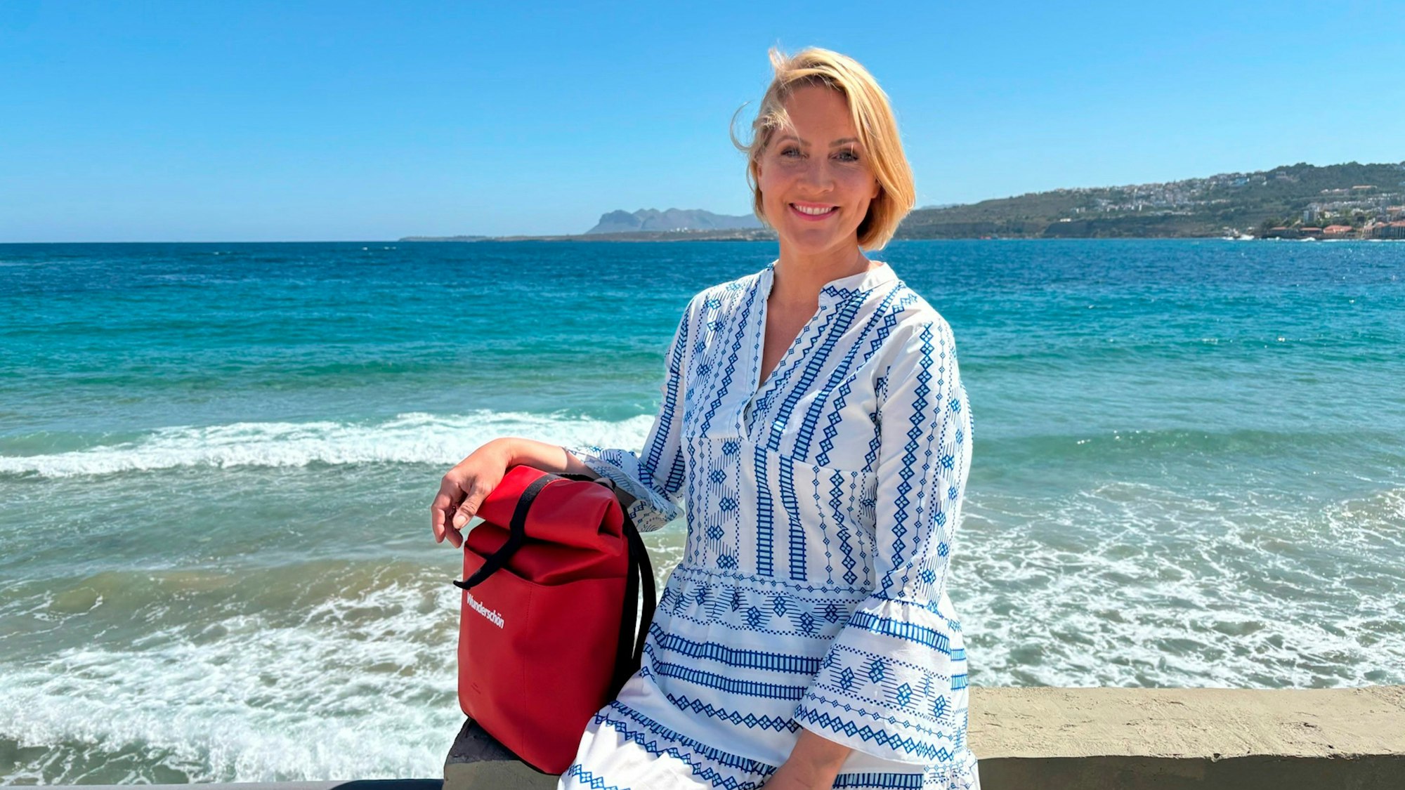 Eine blonde Frau in einem blau-weißen Kleid sitzt auf einer Mauer am Meer