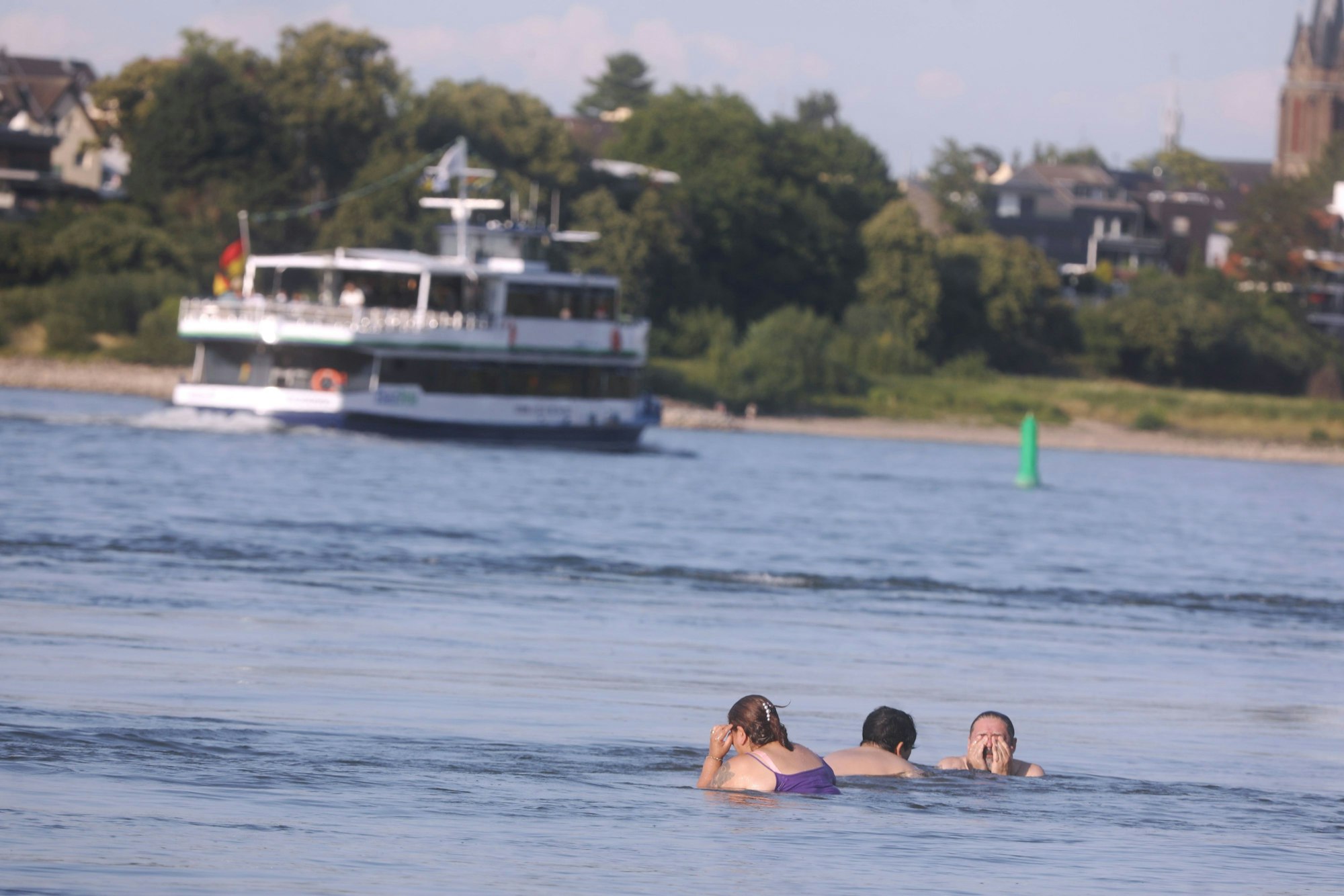 Zwei Frauen und ein Mann baden im Rhein, während ein Schiff vorbeifährt.