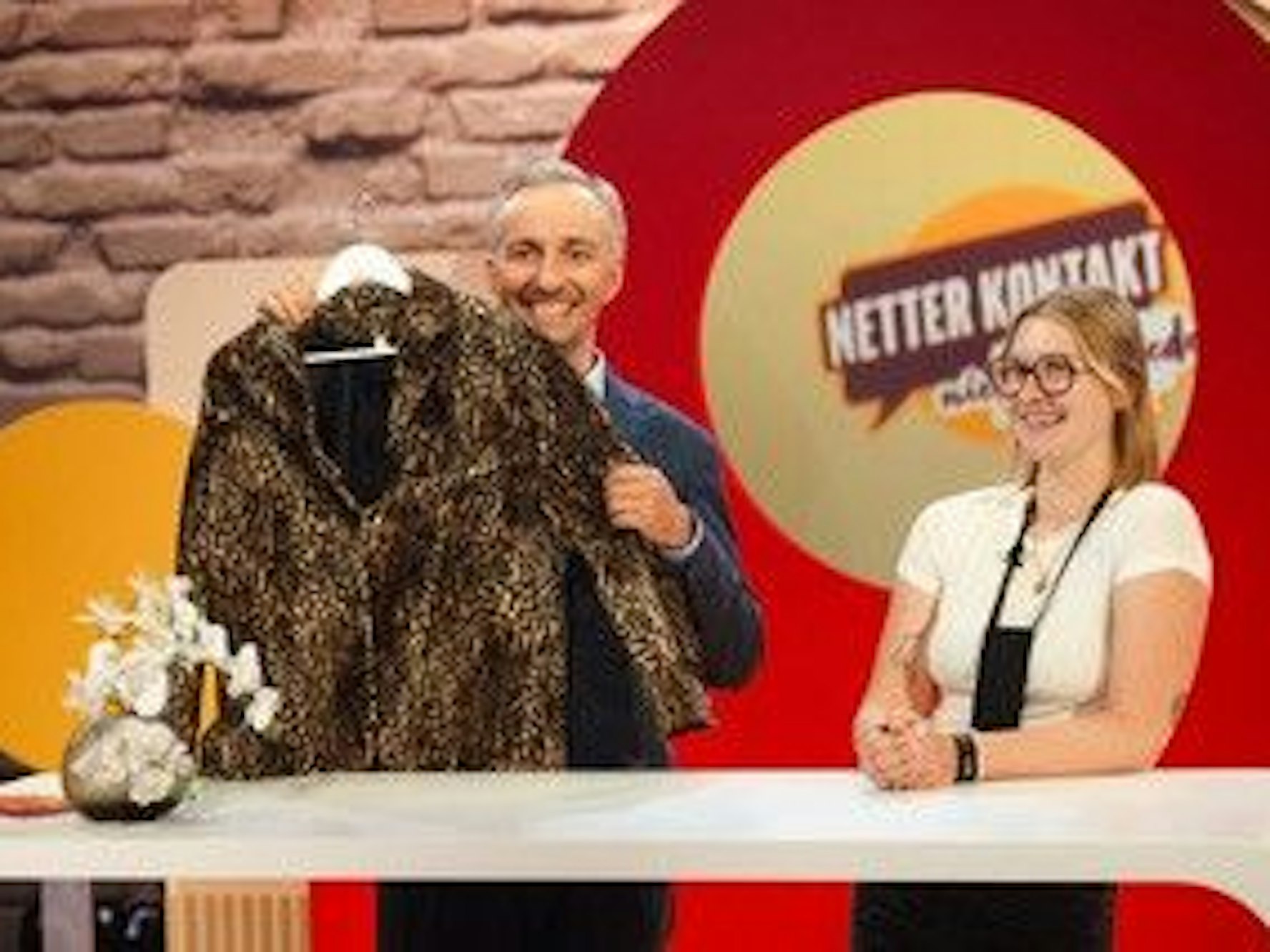 Jan Böhmermann hält eine Jacke in die Kamera, die Kandidatin neben ihm lacht.
