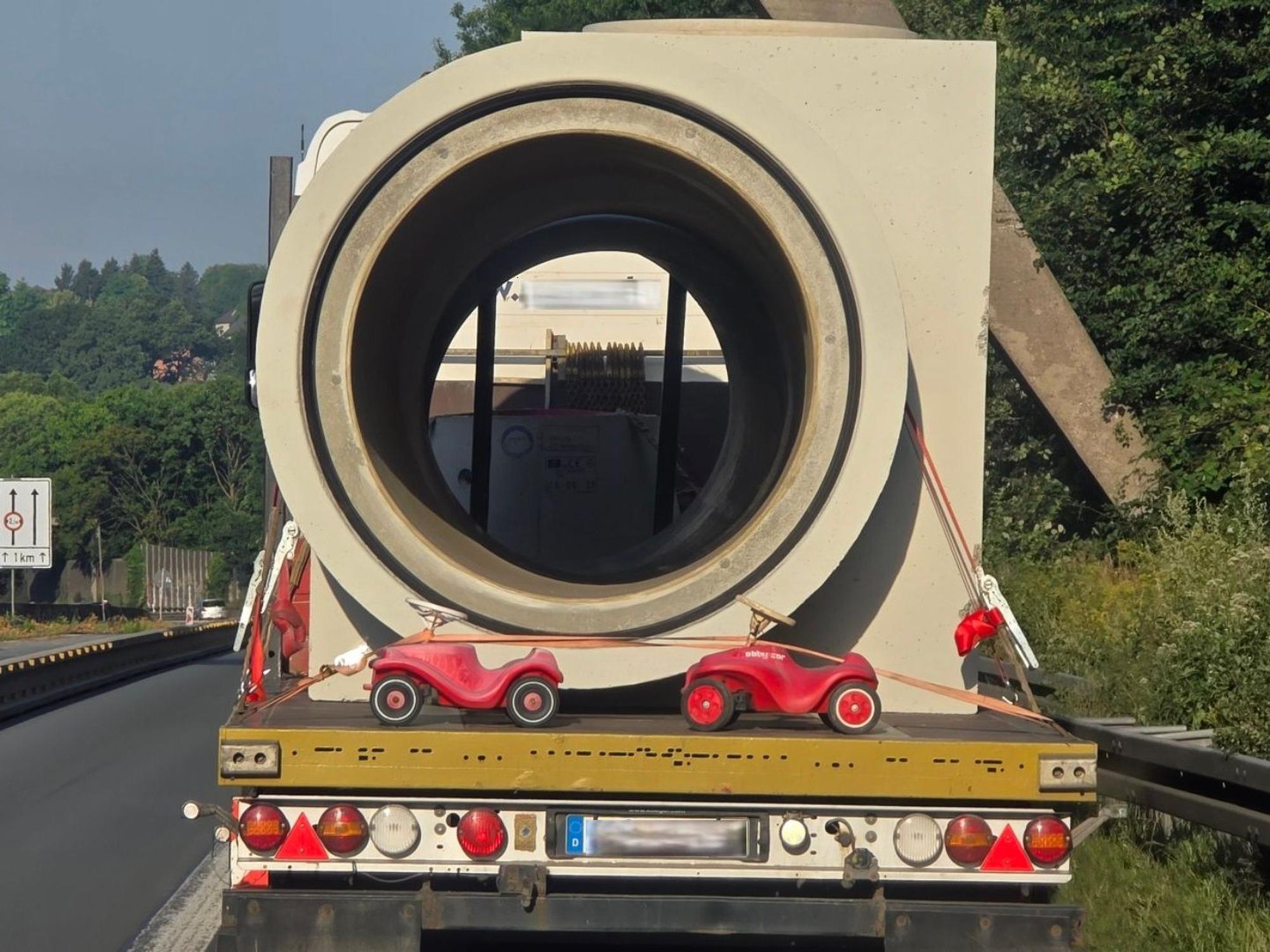Auf einem Transporter, der ein riesiges Betonrohr transportiert, sind zwei kleine Bobbycars festgeschnallt.