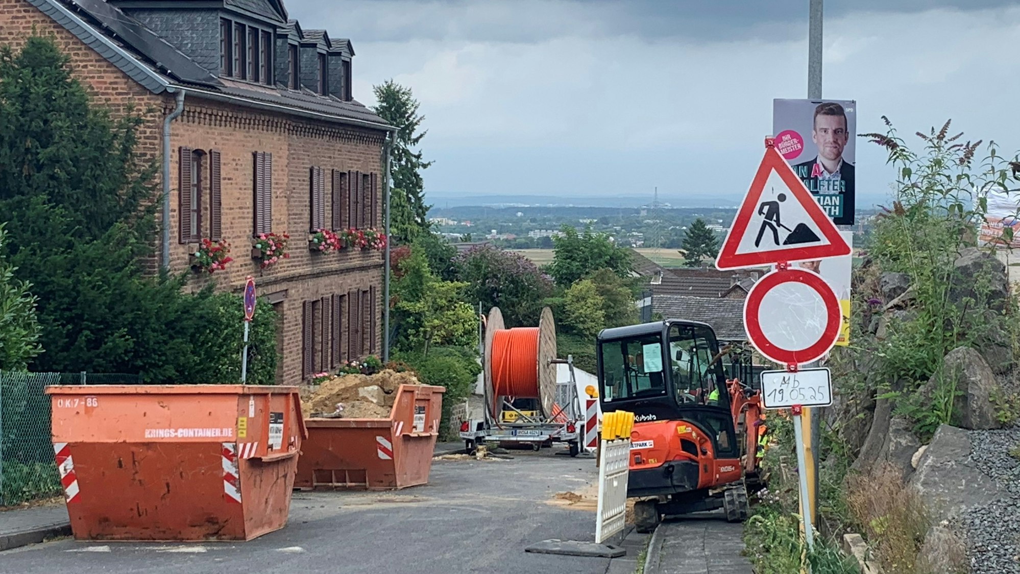 Eine Baustelle mit Absperrungen, Baggern und Schuttcontainern an einer Straße am Hang.