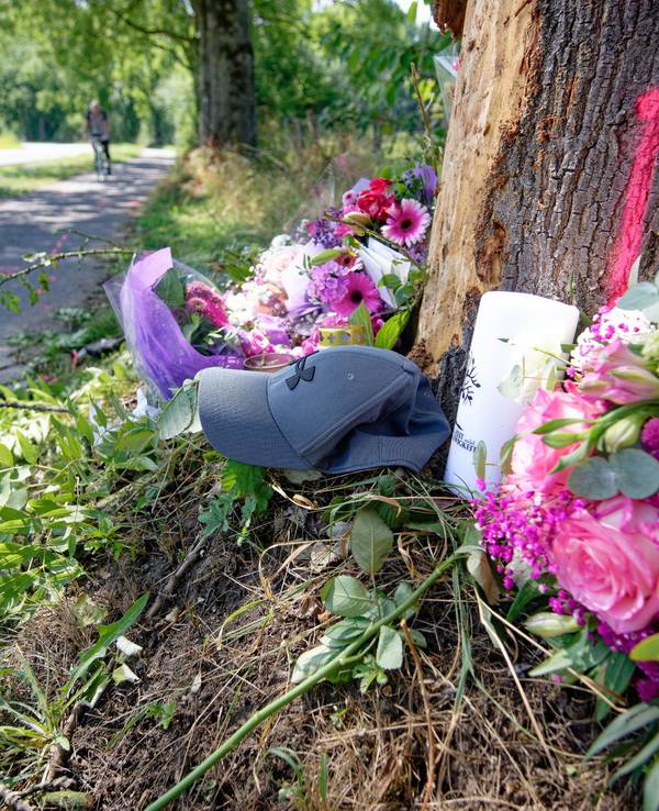 An einem Baumstamm neben einer Straße liegen Blumensträuße und eine Mütze.