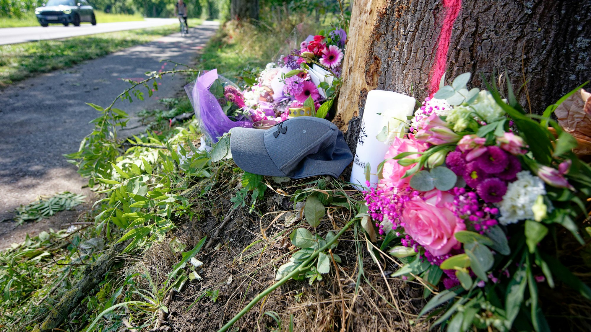 An einem Baumstamm neben einer Straße liegen Blumensträuße und eine Mütze.