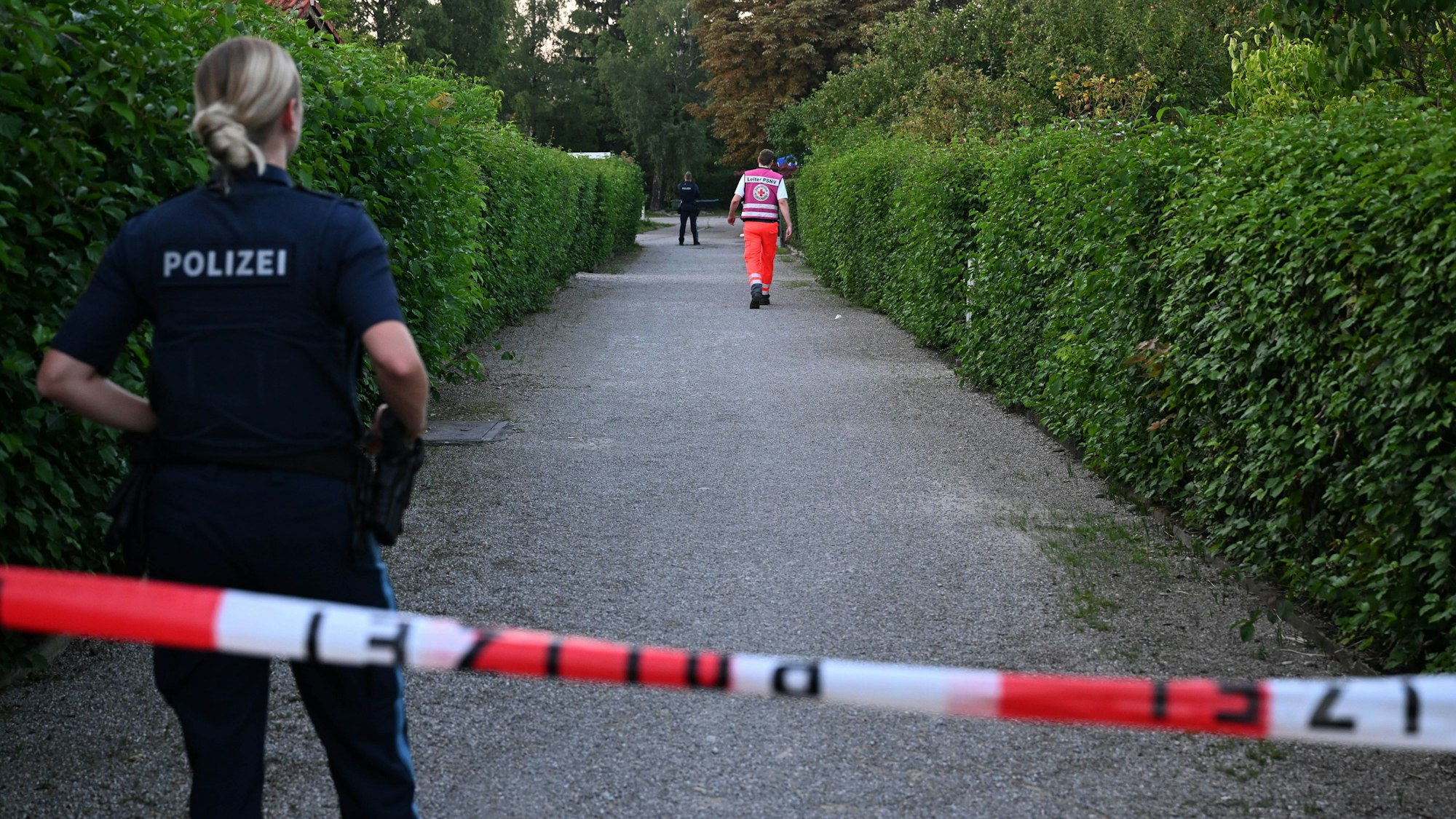 Die Polizei hat einen Gang in einer Kleingartenanlage abgesperrt. Hinter dem Flatterband steht eine Polizistin, weiter entfernt gegen ein Mann vom Rettungsdienst.