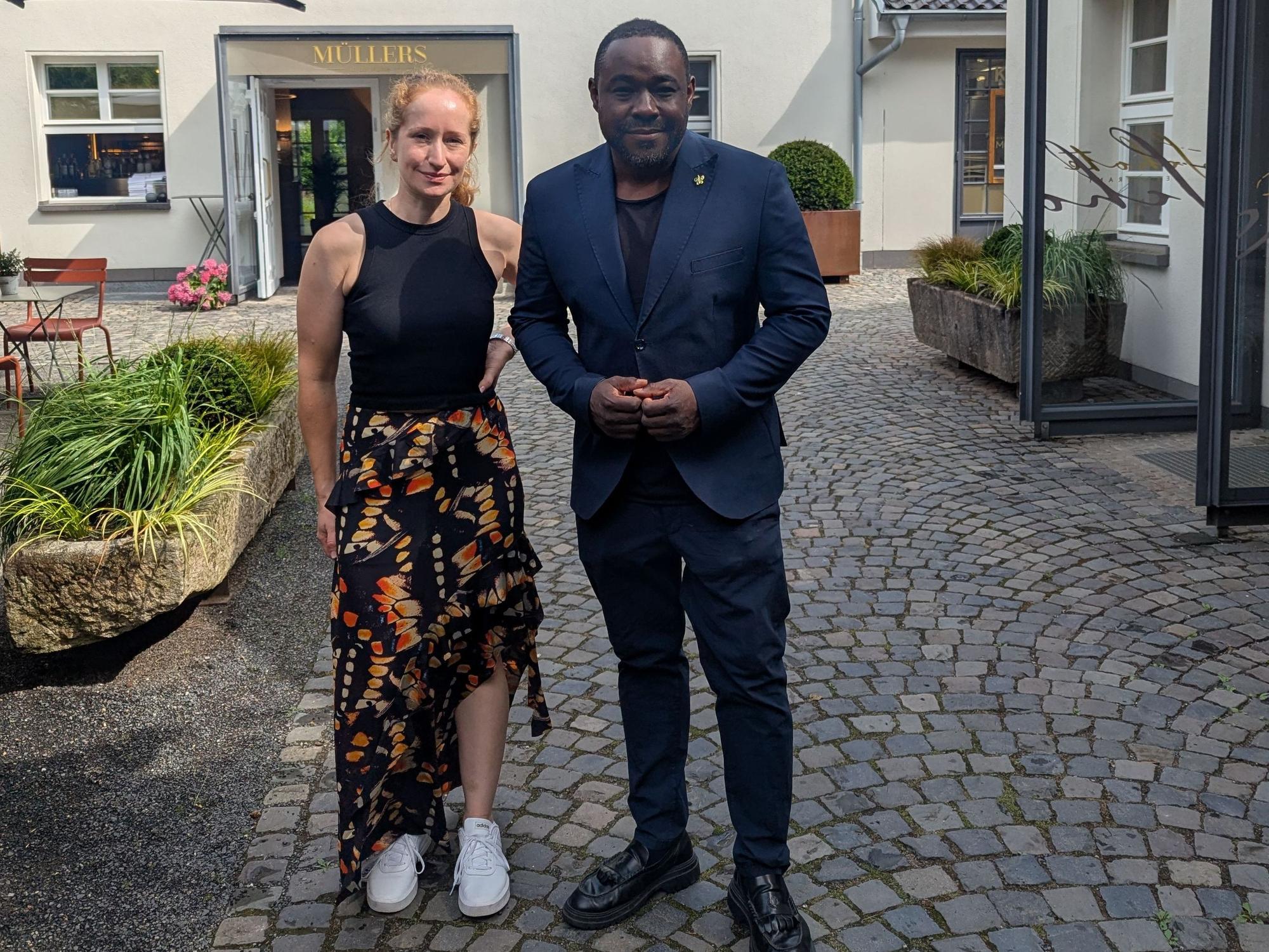 Eine Frau in Sommerrock und schwarzem Tanktop und ein Mann im blauen Anzug stehen auf dem Kopfsteinpflaster vor einem Hotel und lächeln.