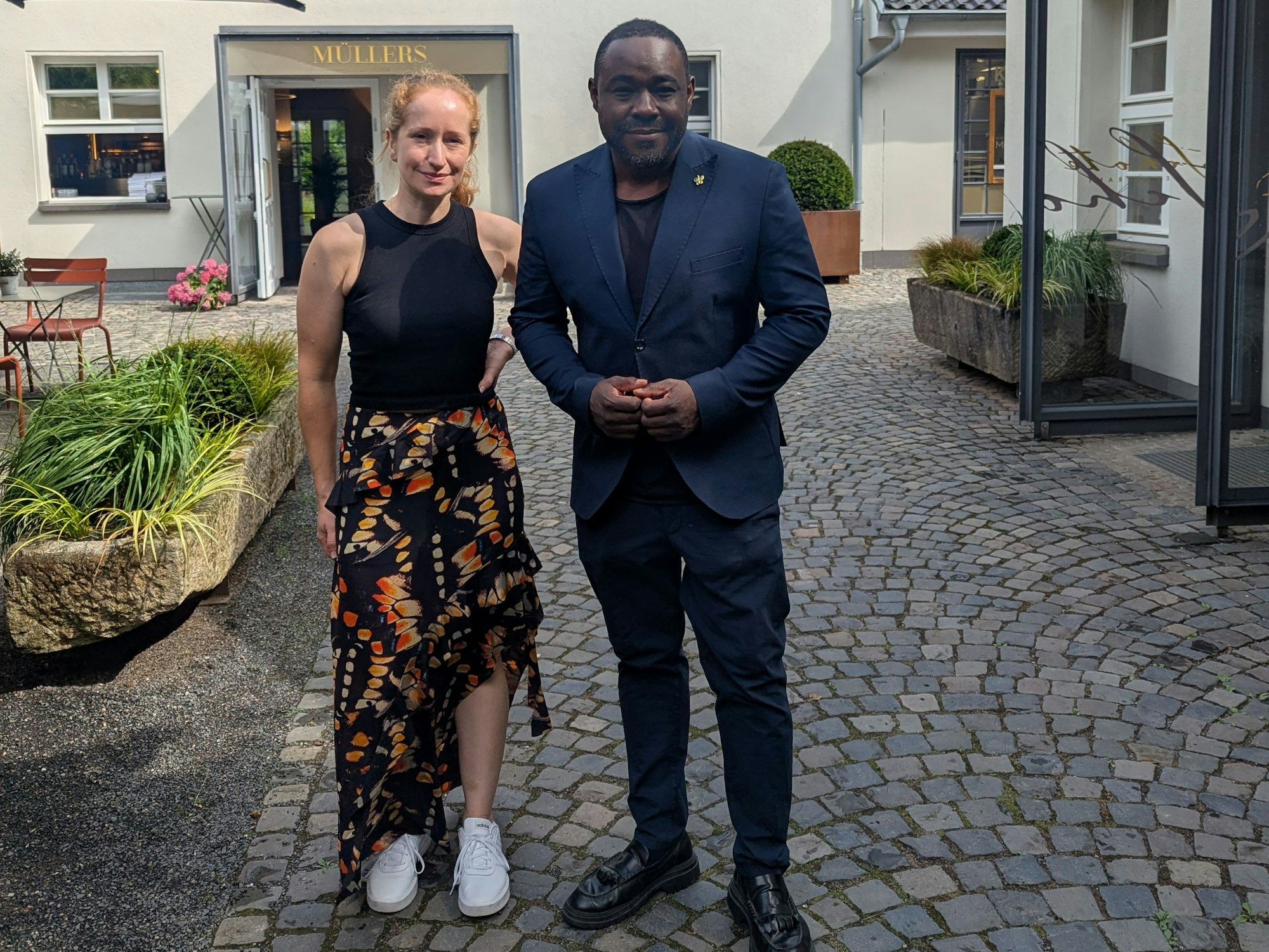 Eine Frau in Sommerrock und schwarzem Tanktop und ein Mann im blauen Anzug stehen auf dem Kopfsteinpflaster vor einem Hotel und lächeln.