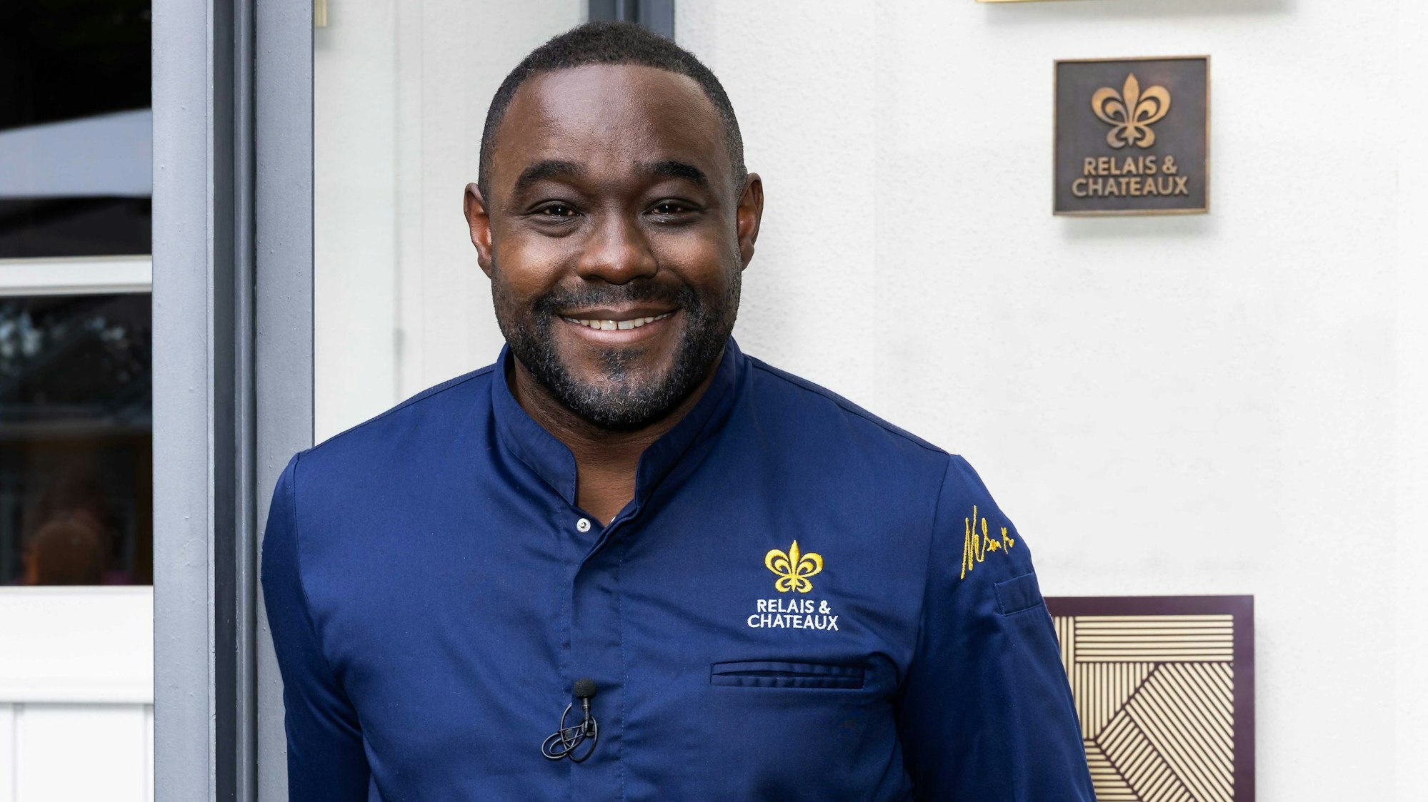 Ein Profikoch in blauer Kochjacke lächelt vor dem Eingang zu einem Hotel