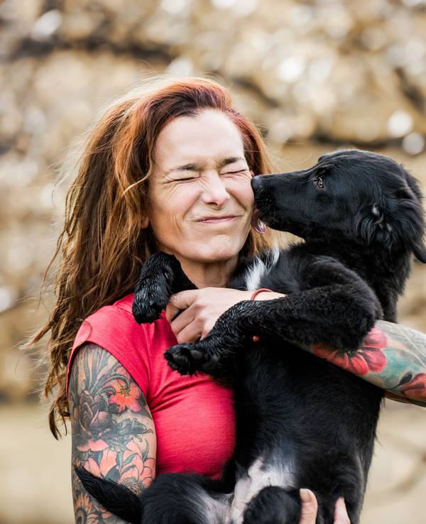 Eine tätowierte Frau hält einen jungen Hund auf dem Arm, der ihr fröhlich durch das Gesicht leckt.