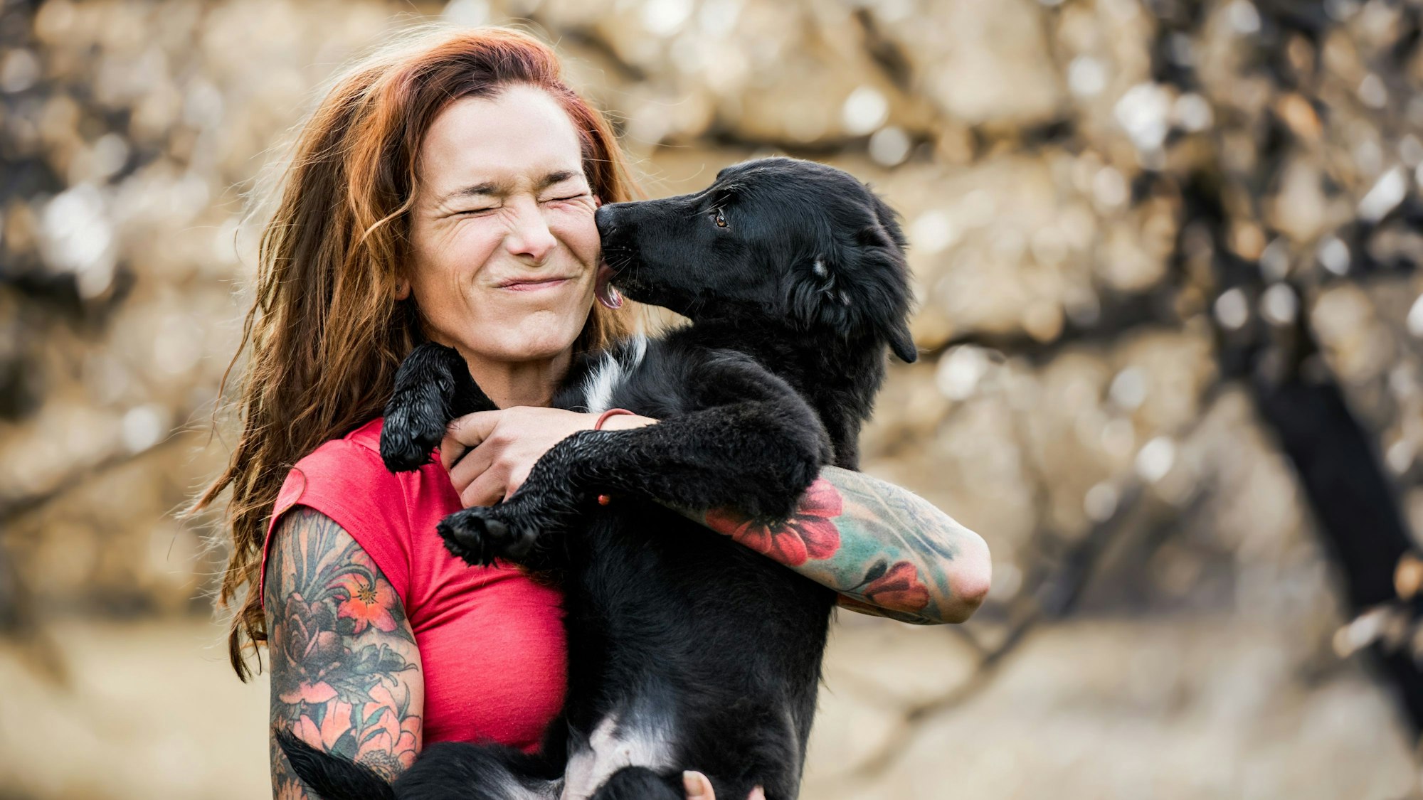 Eine tätowierte Frau hält einen jungen Hund auf dem Arm, der ihr fröhlich durch das Gesicht leckt.