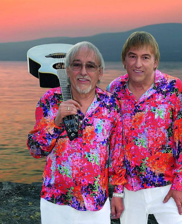 Zwei Männer in kreischbunten Hemden stehen vor einem Sonnenuntergang am Meer. Einer der beiden hat eine weiße Gitarre auf der Schulter