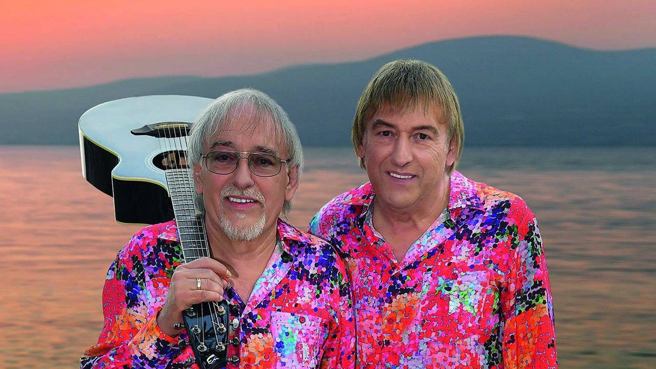 Zwei Männer in kreischbunten Hemden stehen vor einem Sonnenuntergang am Meer. Einer der beiden hat eine weiße Gitarre auf der Schulter