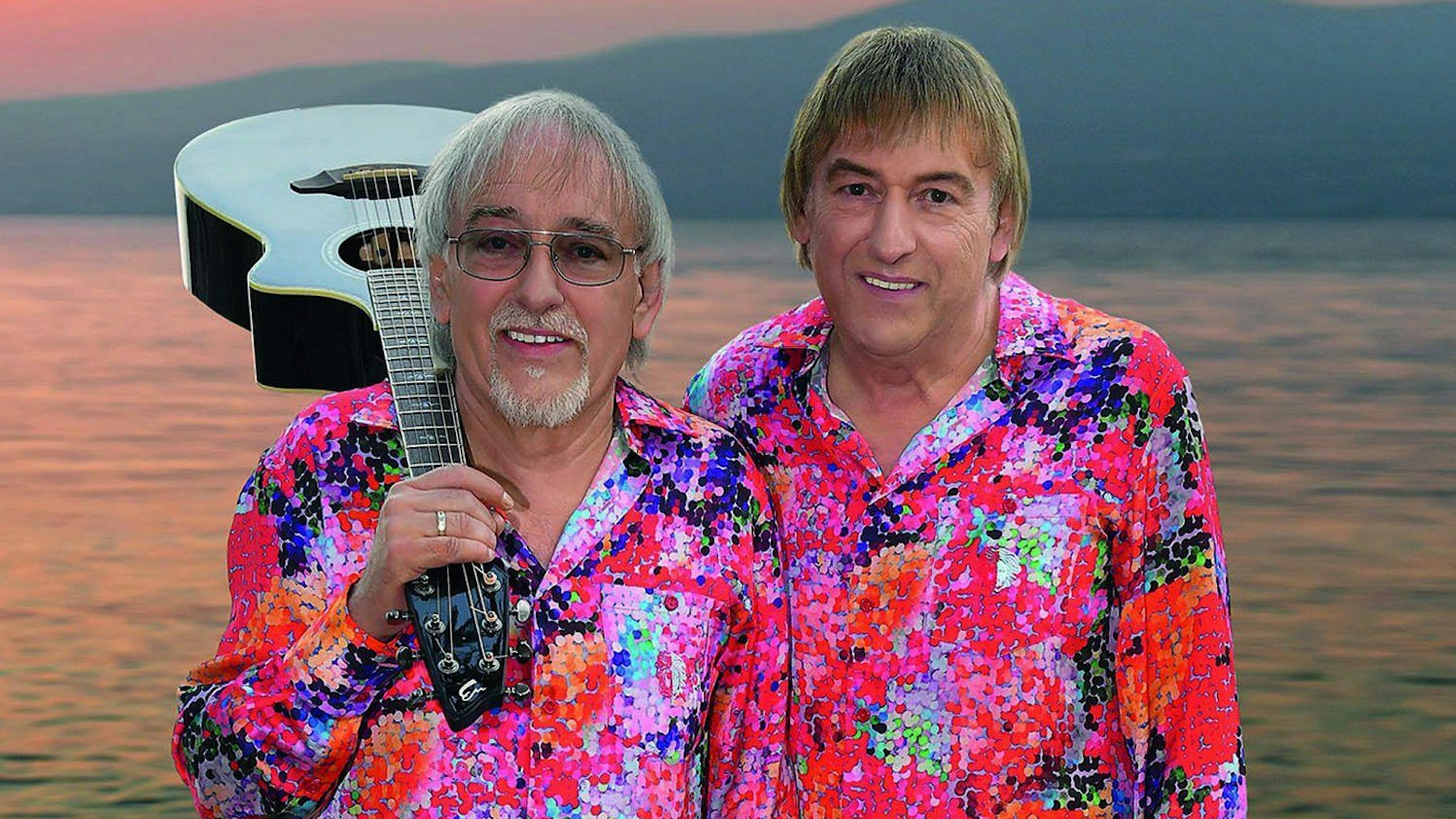 Zwei Männer in kreischbunten Hemden stehen vor einem Sonnenuntergang am Meer. Einer der beiden hat eine weiße Gitarre auf der Schulter