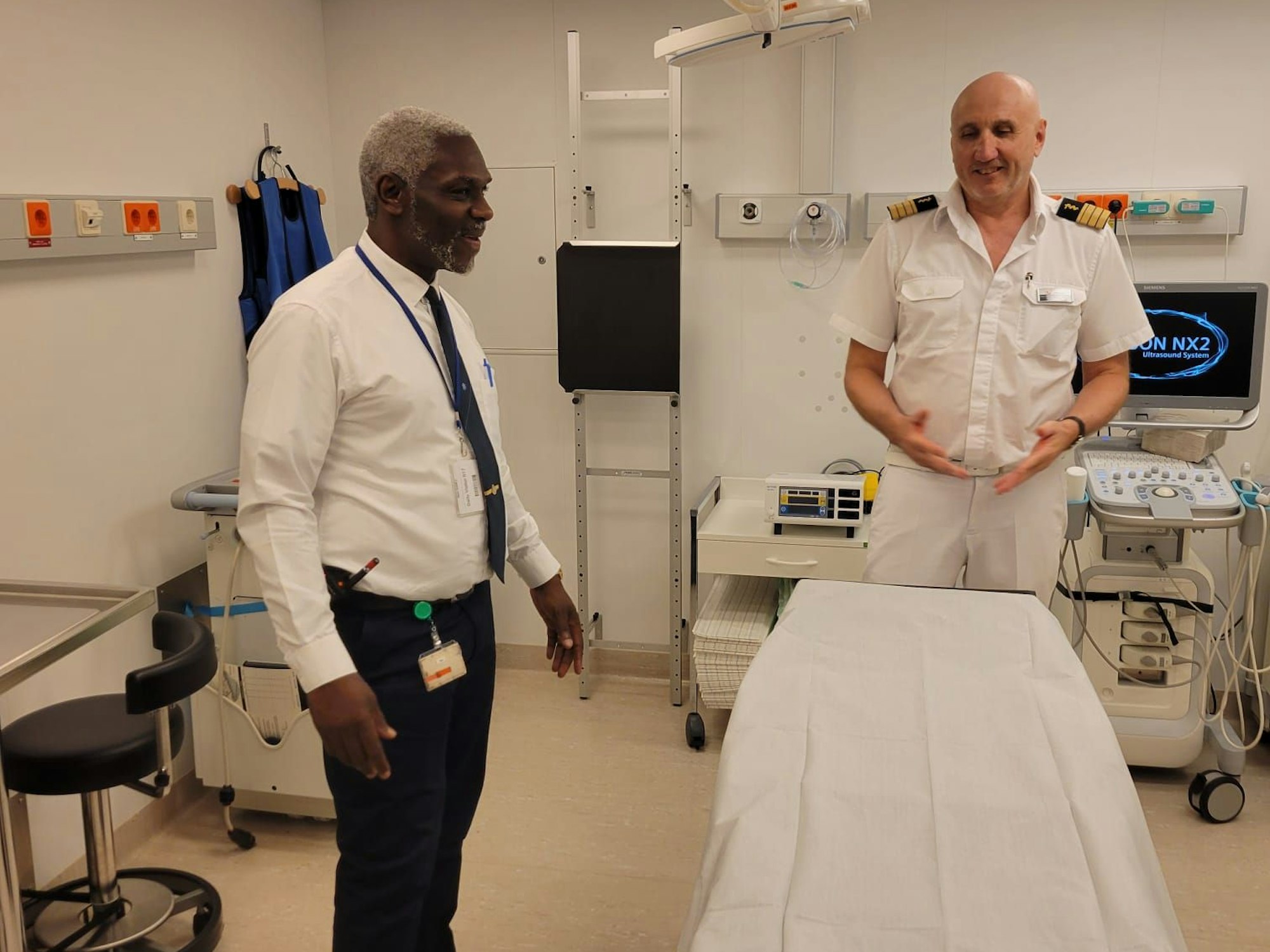Ein Mitarbeiter eines Kreuzfahrtschiffes und ein Schiffsarzt in weißer Uniform stehen in einem kleinen Behandlungsraum vor einer Liege