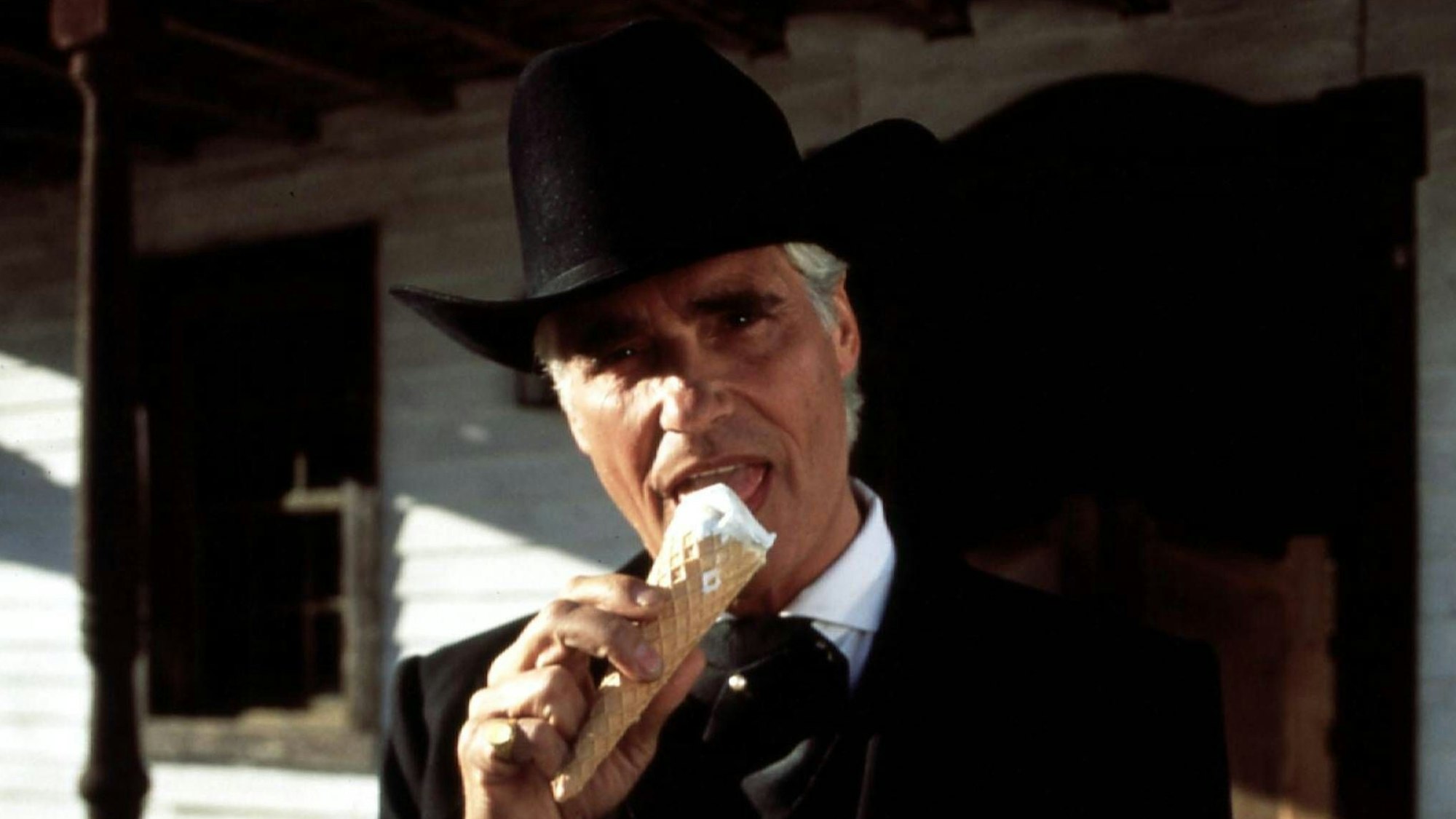 Ein Mann in schwarzer Jacke und mit schwarzem Cowboyhut leckt an einem Eis