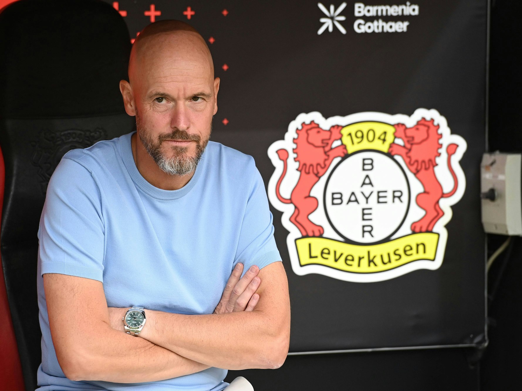 Erik ten Hag sitzt mit verschränkten Armen neben einem Logo von Bayer Leverkusen.