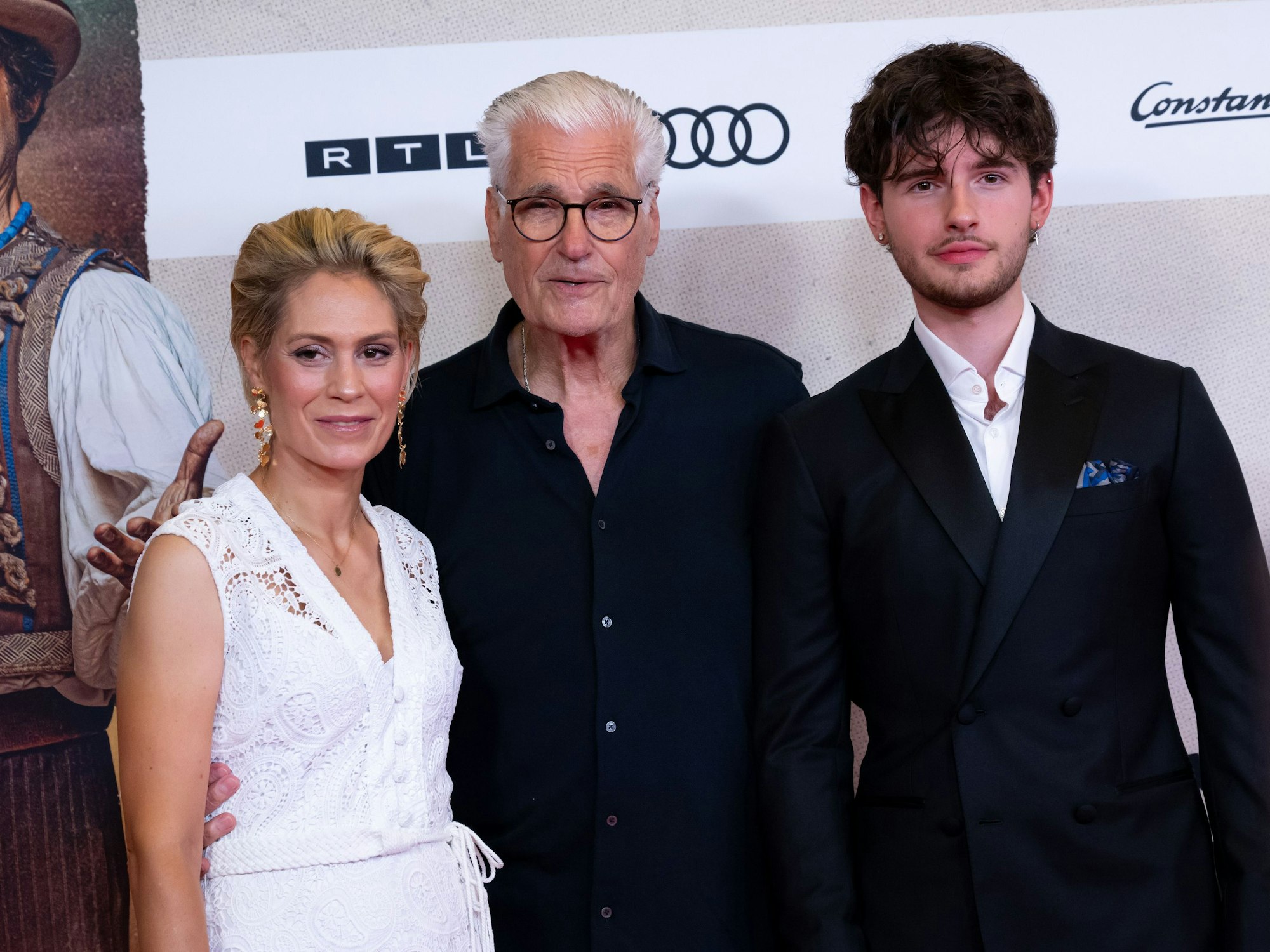 Drei Personen - eine Frau mit Kurzhaarschnitt und in weißem Kleid, ein Mann im dunkeln Hemd mit weißem Haar und ein junger Mann mit Drei-Tage-Bart und Ohrringen stehen für ein Foto zusammen.