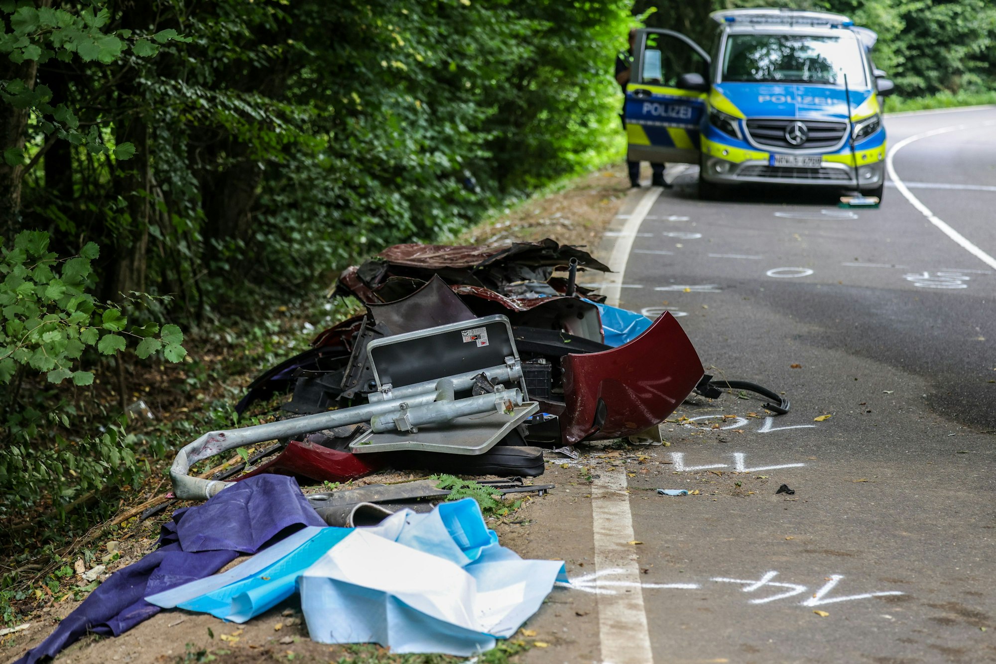 16.08.2025, Nordrhein-Westfalen, Kürten: Trümmerteile liegen am Straßenrand an der Unfallstelle, im Hintergrund ein Fahrzeug der Polizei. Bei einem schweren Autounfall sind am frühen Morgen vier junge Mitfahrer ums Leben gekommen. Der 16-jährige Fahrer sei schwer verletzt worden, sagte eine Sprecherin des Rheinisch-Bergischen Kreises. Foto: Christoph Reichwein/dpa +++ dpa-Bildfunk +++