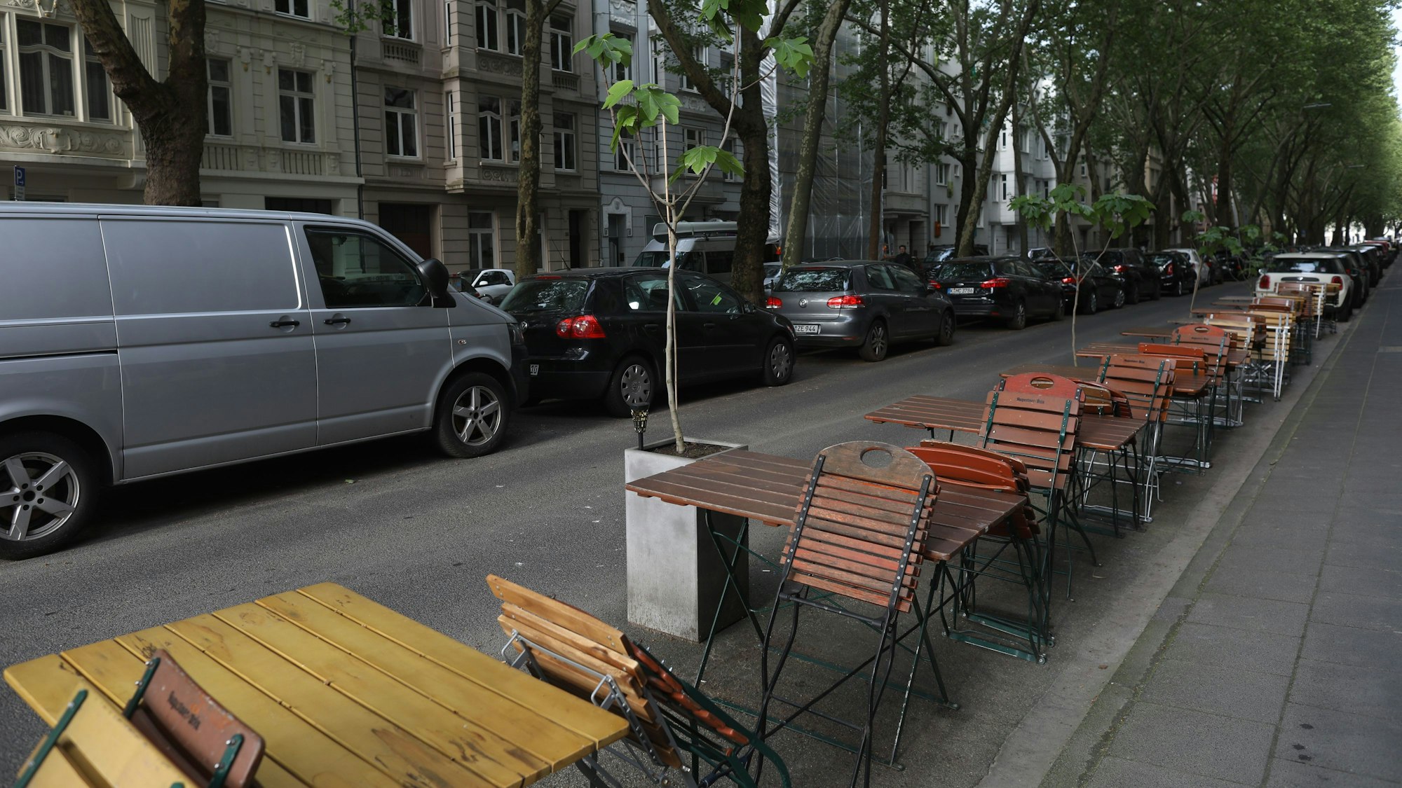 Auf einer Straße parken links dicht an dicht Fahrzeuge, auf der rechten Seite hat ein Lokal Tische und Stühle aufgestellt.