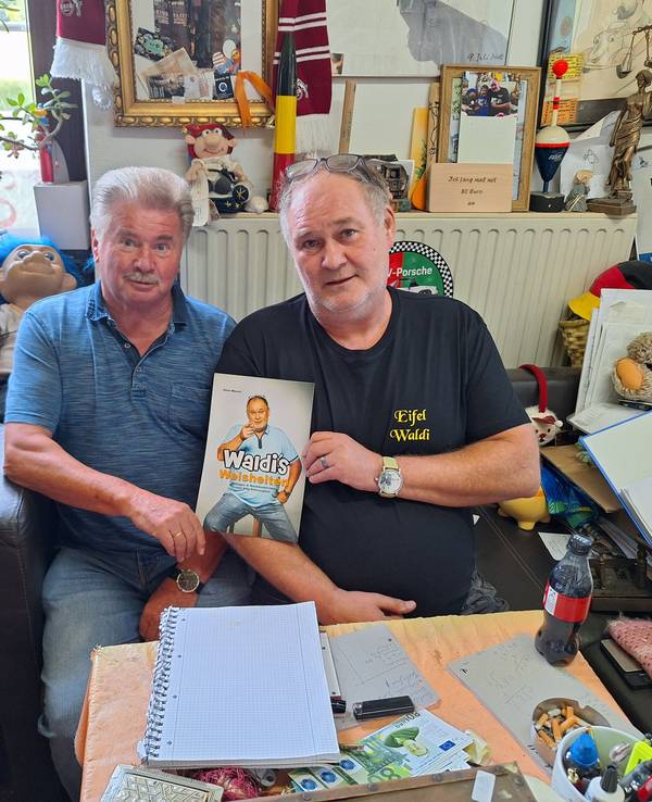 Zwei ältere Männer sitzen vor einer Heizung und halten ein Buch in die Kamera