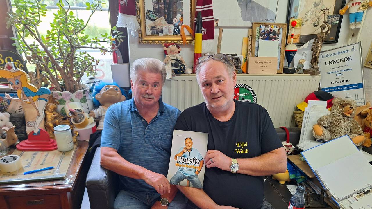 Zwei ältere Männer sitzen vor einer Heizung und halten ein Buch in die Kamera