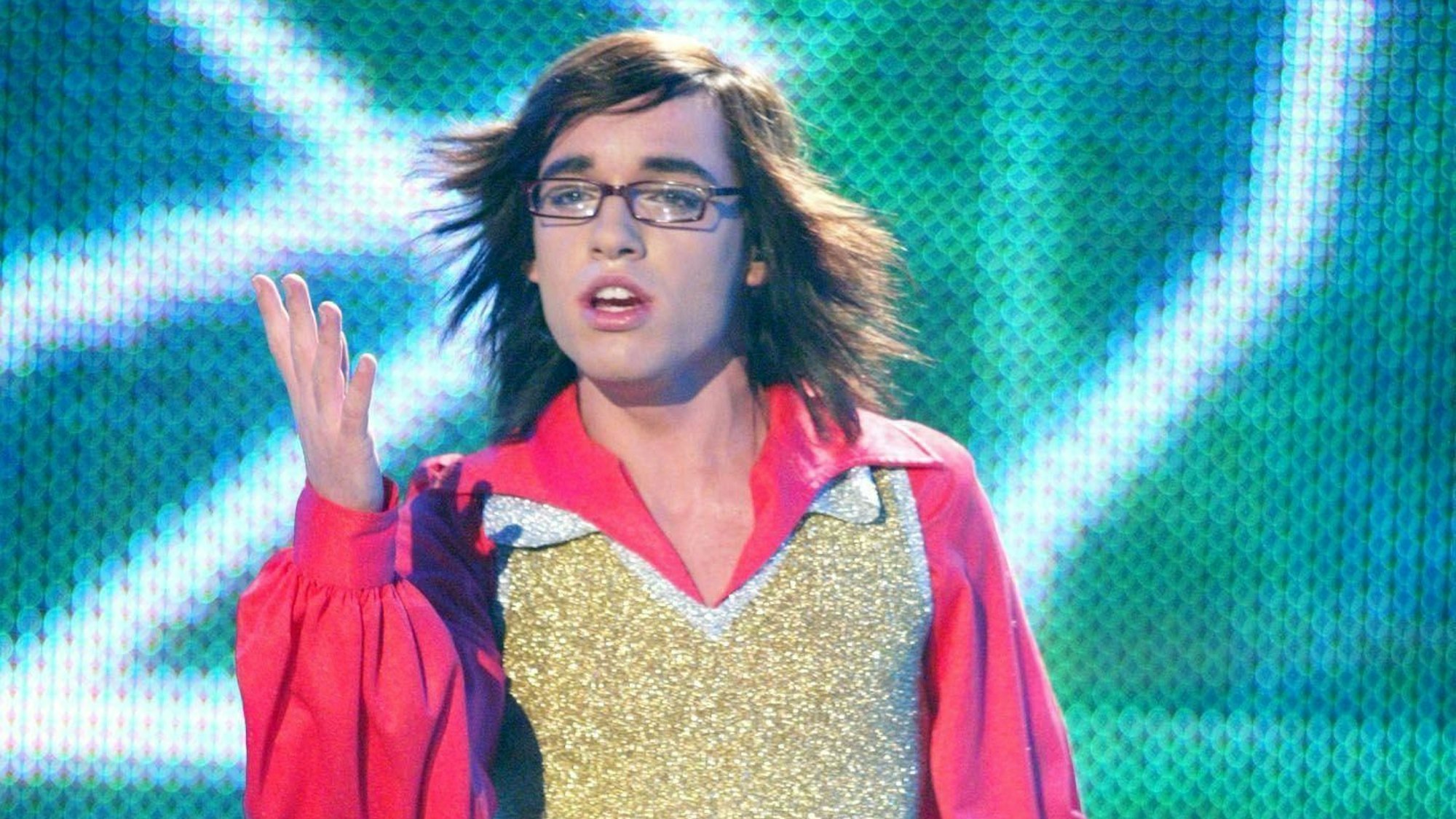 Ein junger Mann mit Brille, Sturmfrisur und Pailettenweste singt auf einer Bühne