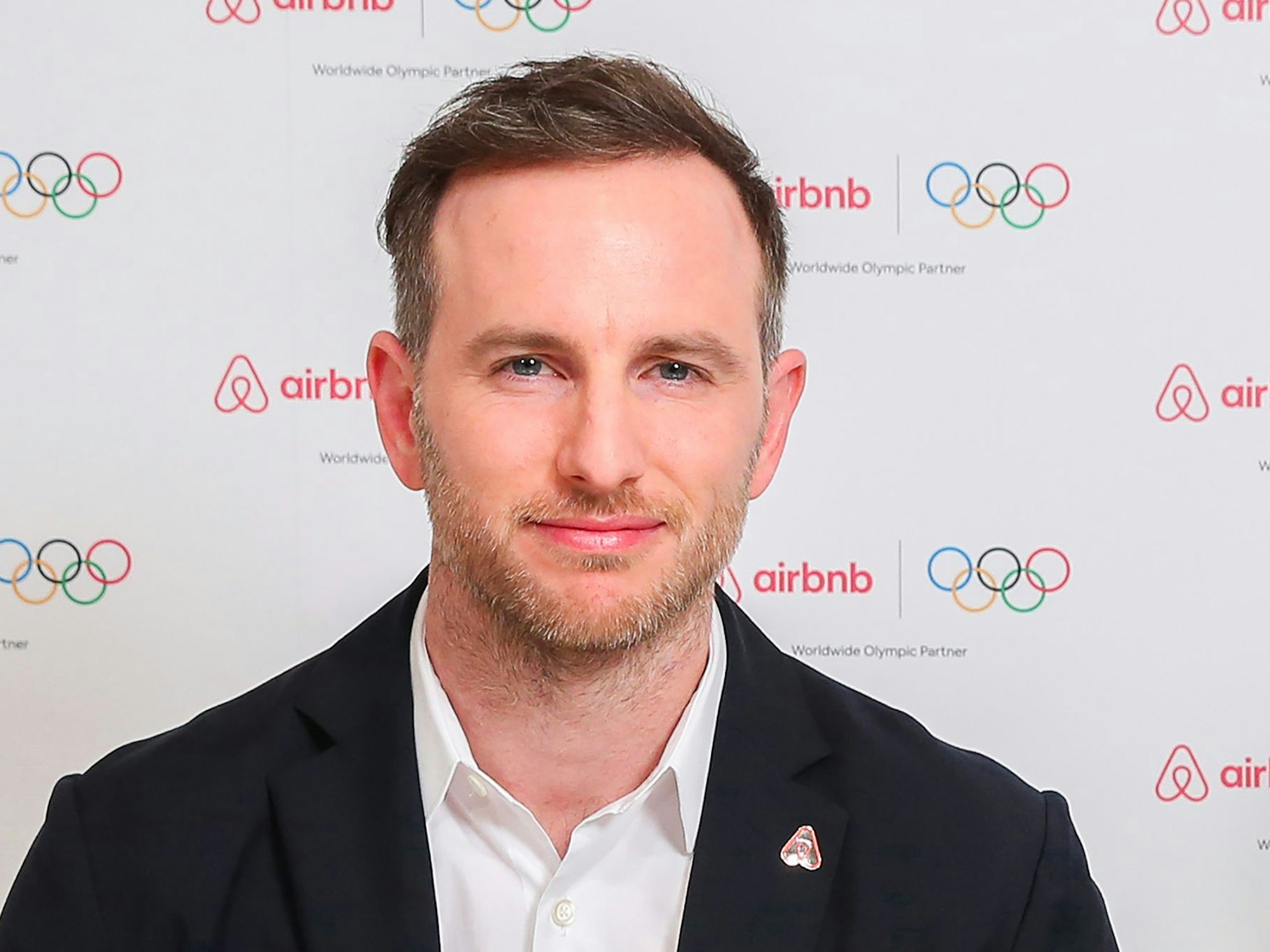 Ein Mann steht vor einer Wand, auf der mehrfach das Wort airbnb und die olympischen Ringe gedruckt sind.