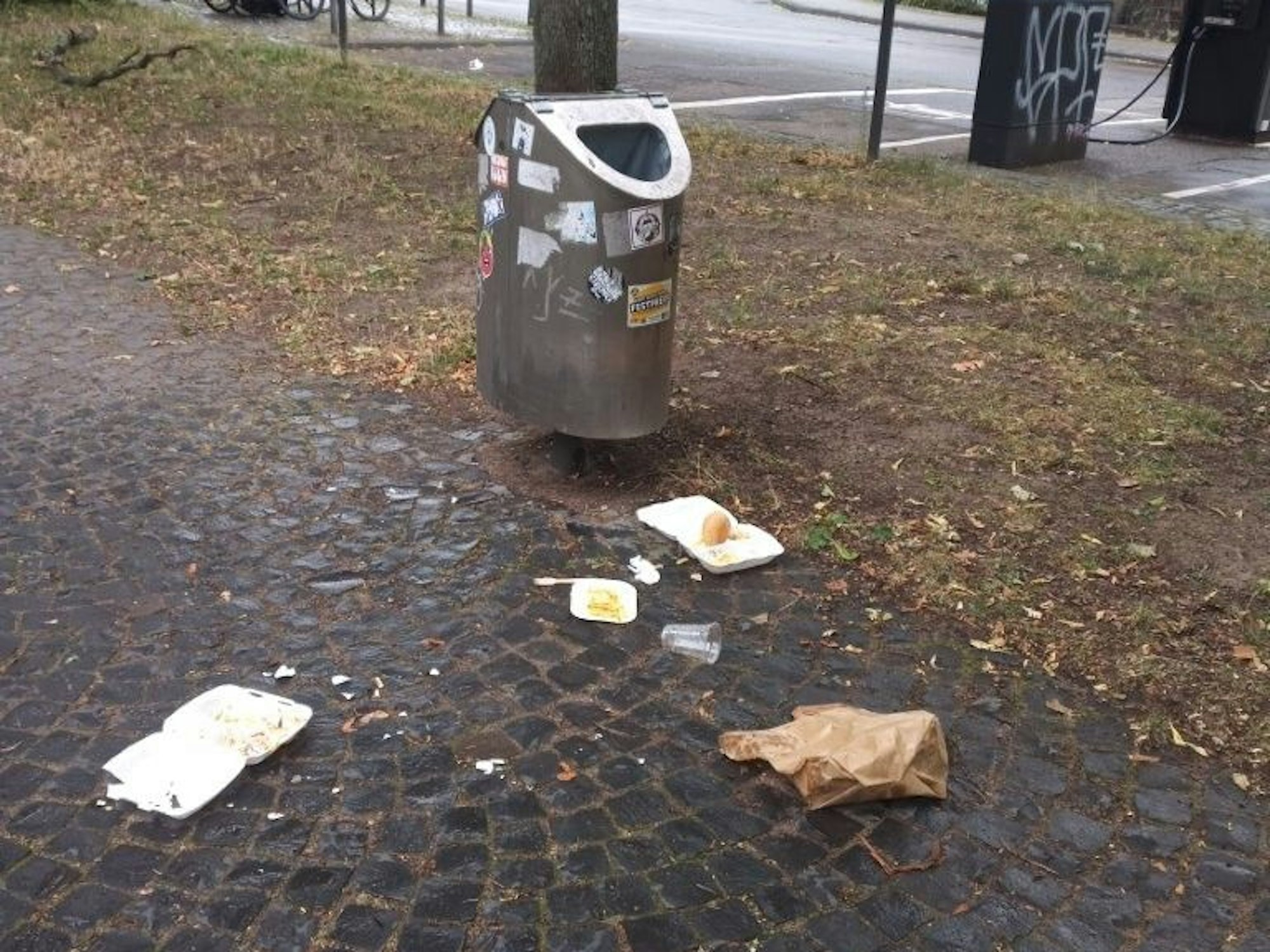 Vor einem leeren Mülleimer liegen Essensverpackungen, zum Teil mit Resten, auf dem Boden.