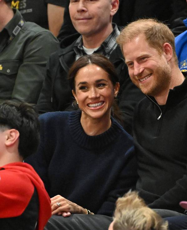 Harry und Meghan posieren auf der Tribüne bei einem Sportevent für ein Foto.
